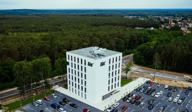Biały, nowoczesny budynek biurowy z logo firmy na elewacji, otoczony parkingiem i lasem, widok z lotu ptaka.