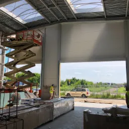 Montaż paneli warstwowych w hali produkcyjnej: pracownicy na podnośniku koszowym mocują panele do konstrukcji stalowej, widoczny otwarty wjazd oraz materiały budowlane.