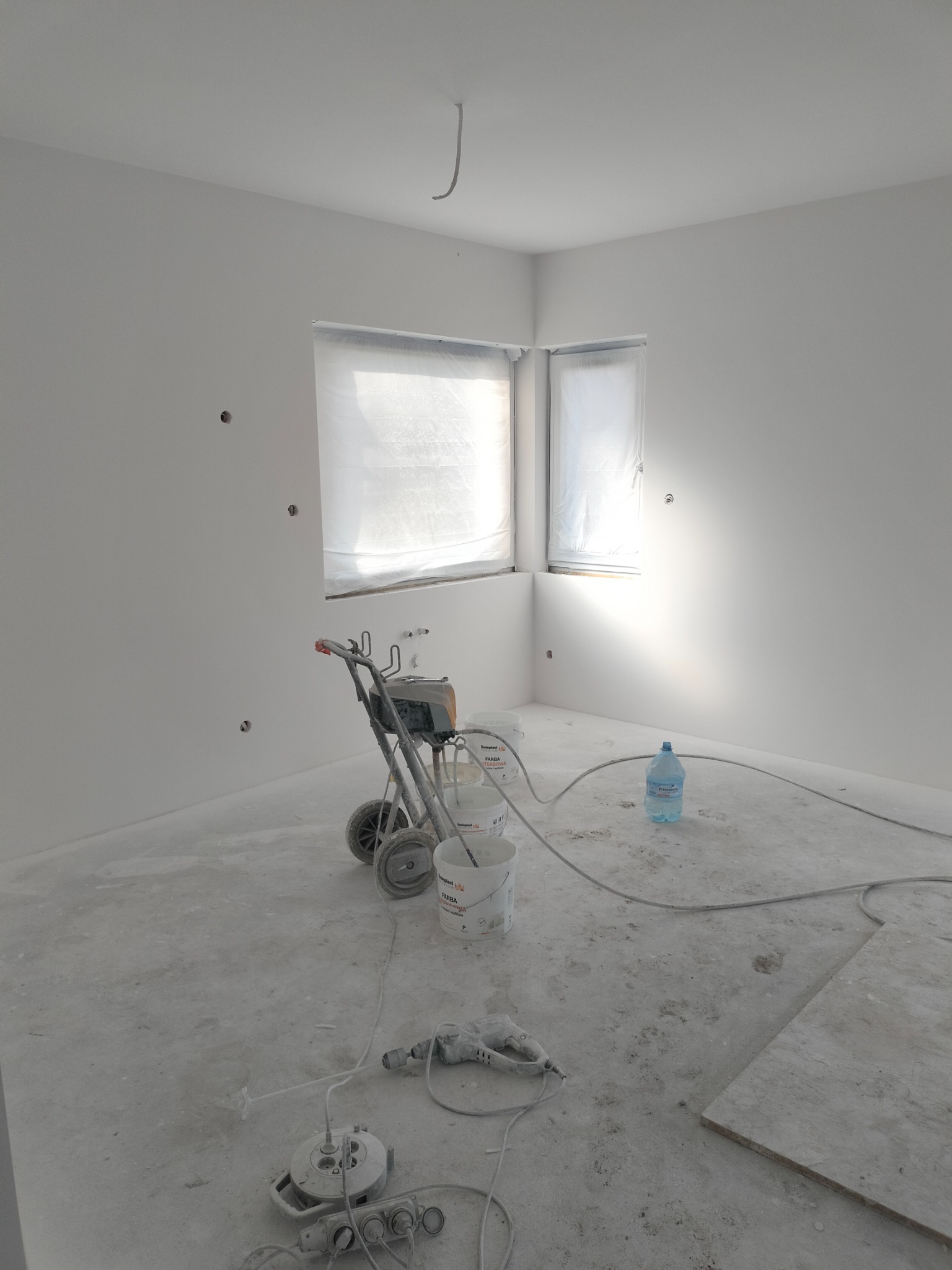 Wnętrze świeżo pomalowanego na biało pokoju z zakrytymi oknami, widoczny agregat malarski, puszki z farbą, przedłużacz i butelka wody na podłodze.