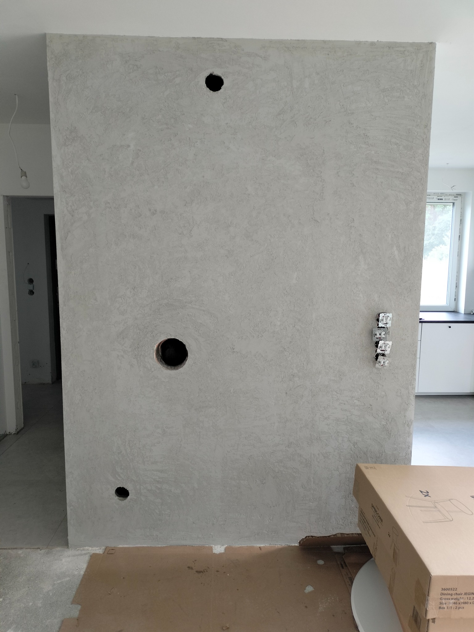 Ściana z surowym, jasnoszarym tynkiem o nieregularnej fakturze, widoczne otwory pod instalacje elektryczne oraz zamontowane puszki z przełącznikami, podłoga zabezpieczona kartonem.