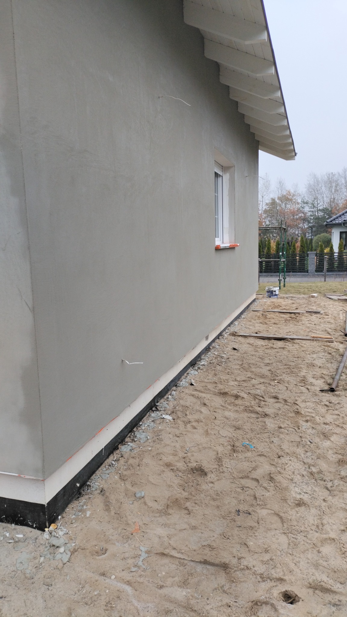 Świeżo nałożony, jasnoszary tynk maszynowy na ścianie domu jednorodzinnego w Gorzowie Wielkopolskim, widok z zewnątrz, z widocznym oknem i elementami izolacji przy gruncie.