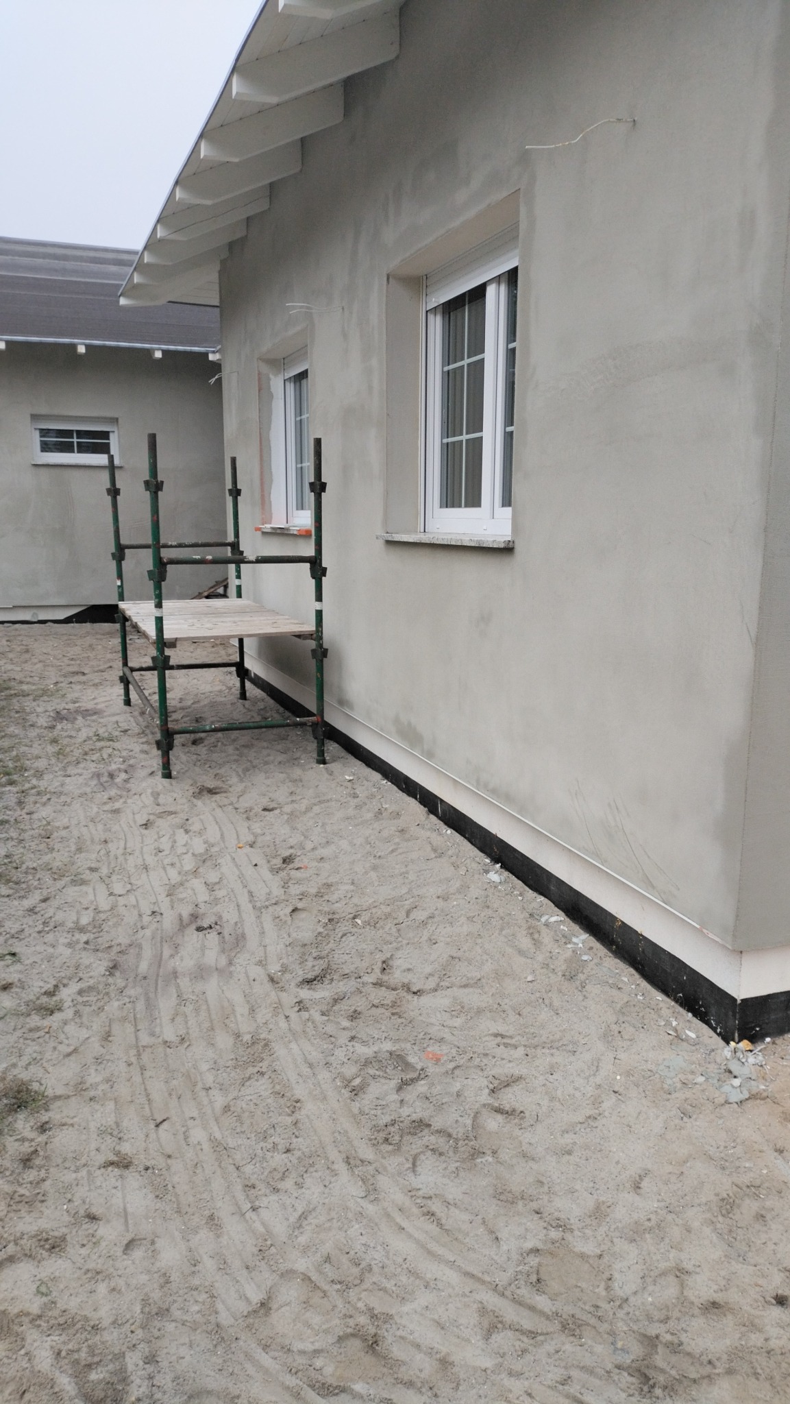 Świeżo otynkowana ściana budynku z białymi oknami, widoczne rusztowanie i piasek na ziemi.