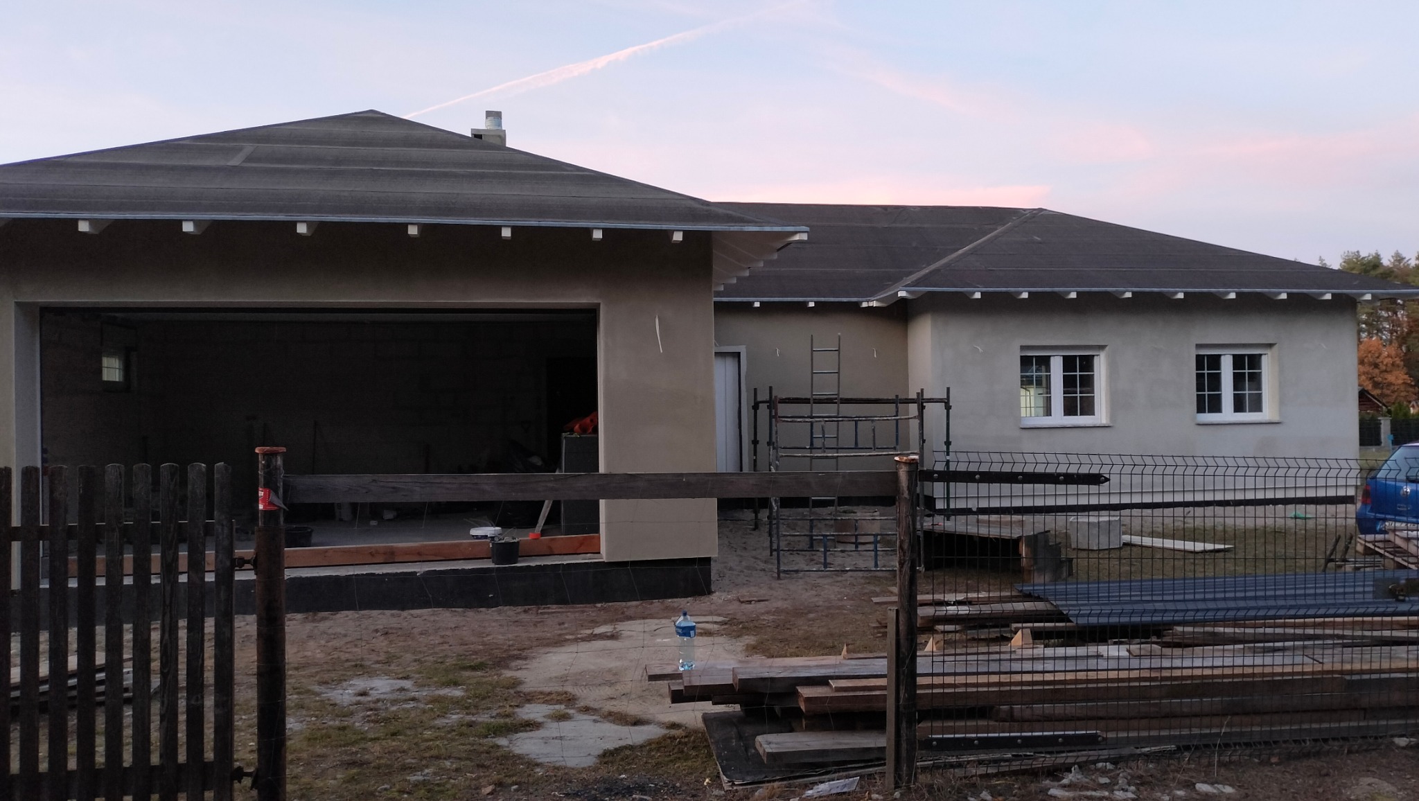 Surowy stan domu jednorodzinnego z garażem po nałożeniu tynku, widoczne rusztowania i przygotowane deski, ogrodzenie z siatki, zachmurzone niebo.