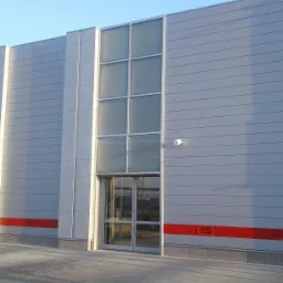 Szara hala przemysłowa z panelami ściennymi, dużymi oknami, czerwonym pasem u dołu i widocznymi elementami instalacji zewnętrznej.