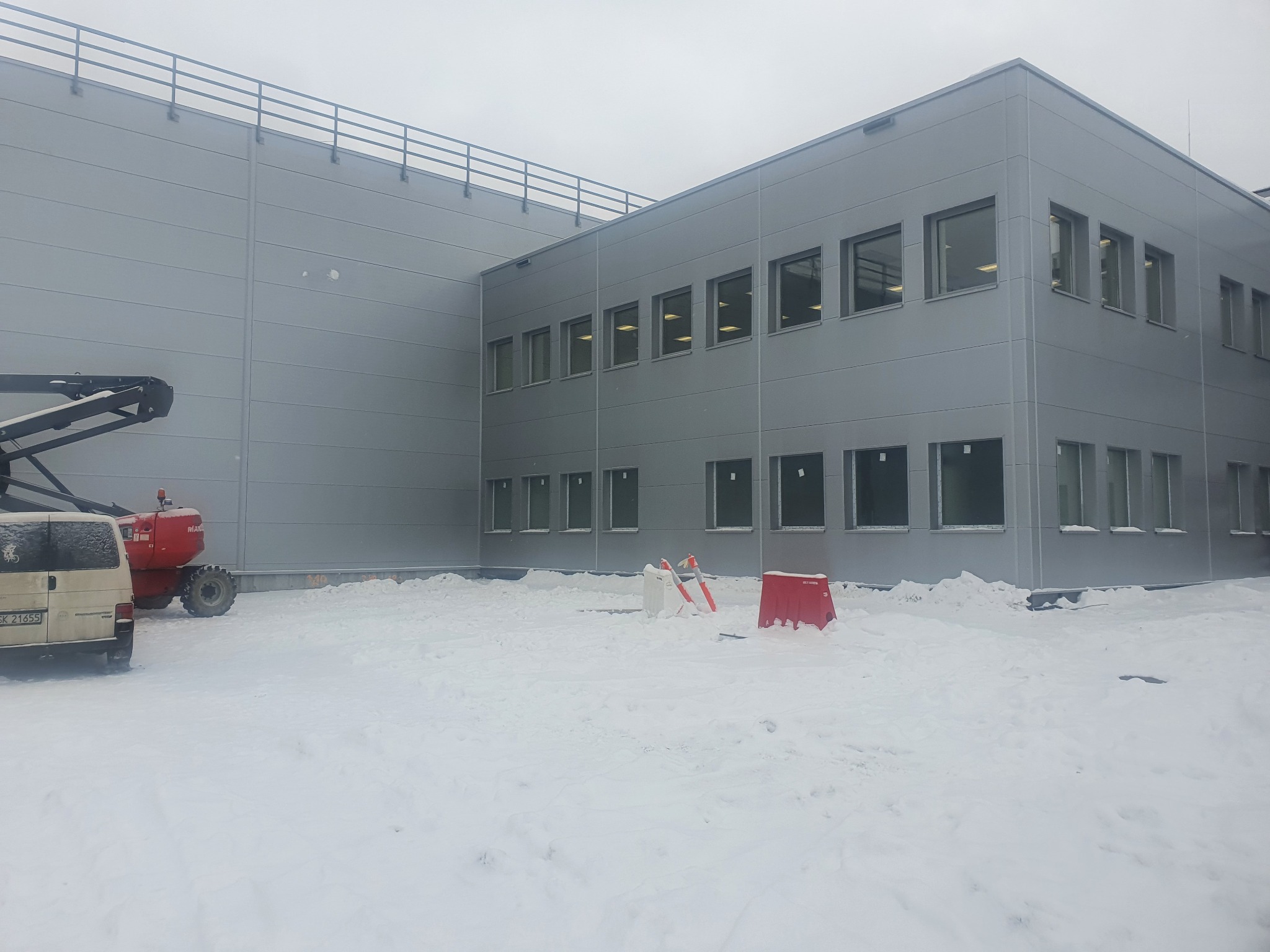 Szary budynek przemysłowy z rzędem okien, widoczny podnośnik koszowy i samochód dostawczy na ośnieżonym terenie.