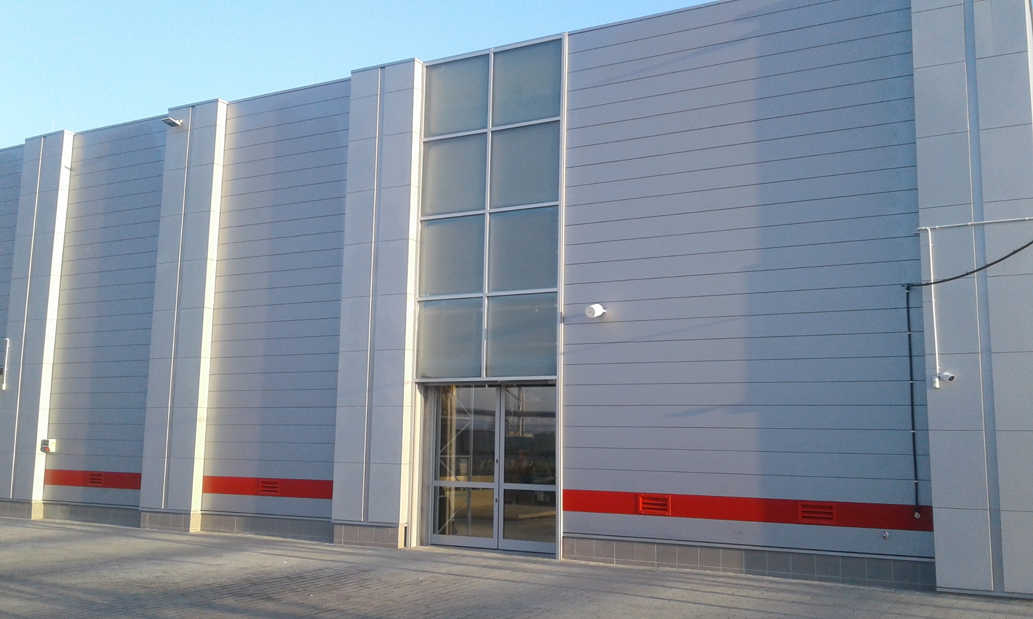 Szara hala przemysłowa z panelami ściennymi, dużymi oknami, czerwonym pasem u dołu i widocznymi elementami instalacji zewnętrznej.
