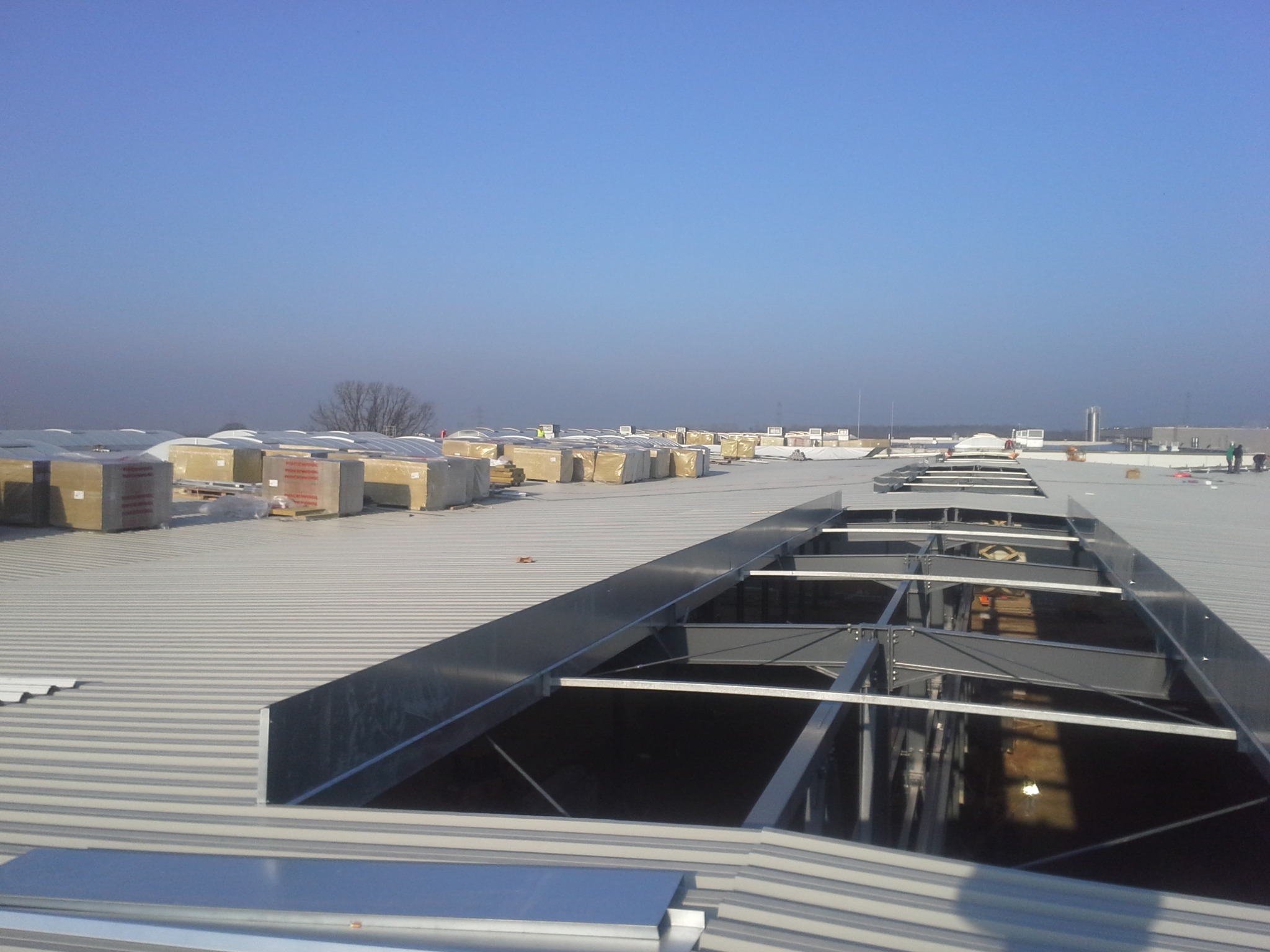 Montaż świetlików dachowych na blaszanym dachu hali przemysłowej, z widocznymi paczkami materiałów izolacyjnych w tle i błękitnym niebem.