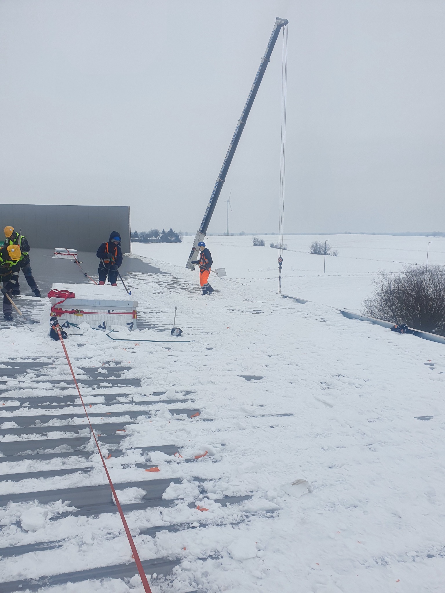 Pracownicy w kombinezonach usuwają śnieg z dachu hali przemysłowej przy użyciu łopat, w tle widoczny dźwig i zasypane śniegiem pole.