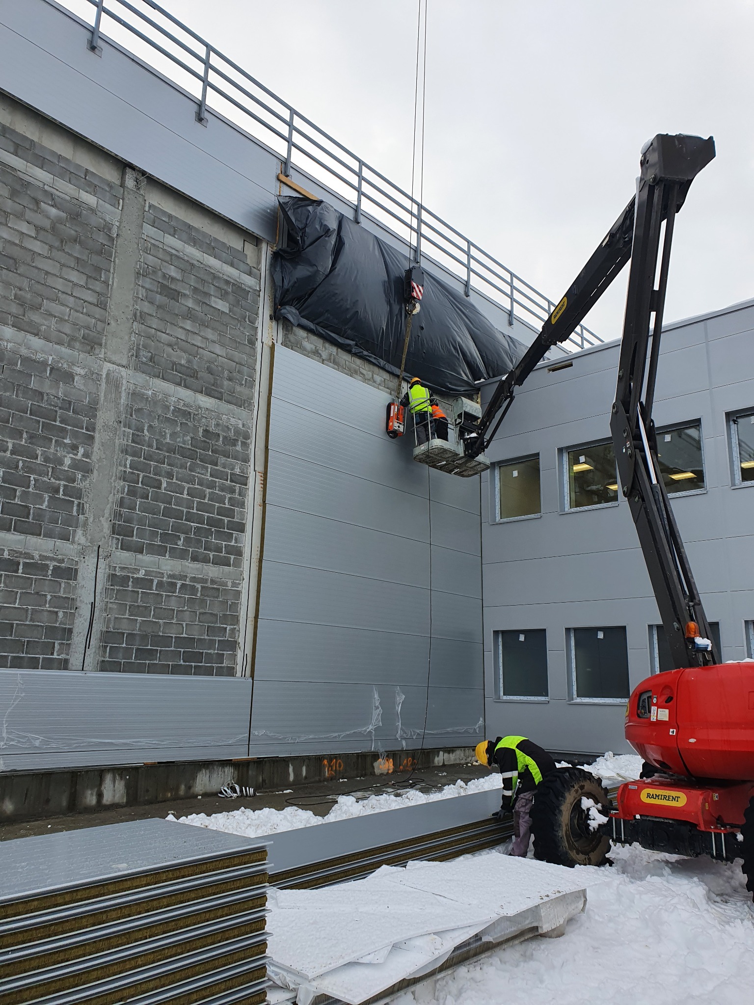 Montaż paneli ściennych na budynku przemysłowym w zimowych warunkach, z użyciem podnośnika koszowego i widoczną stertą paneli na śniegu.