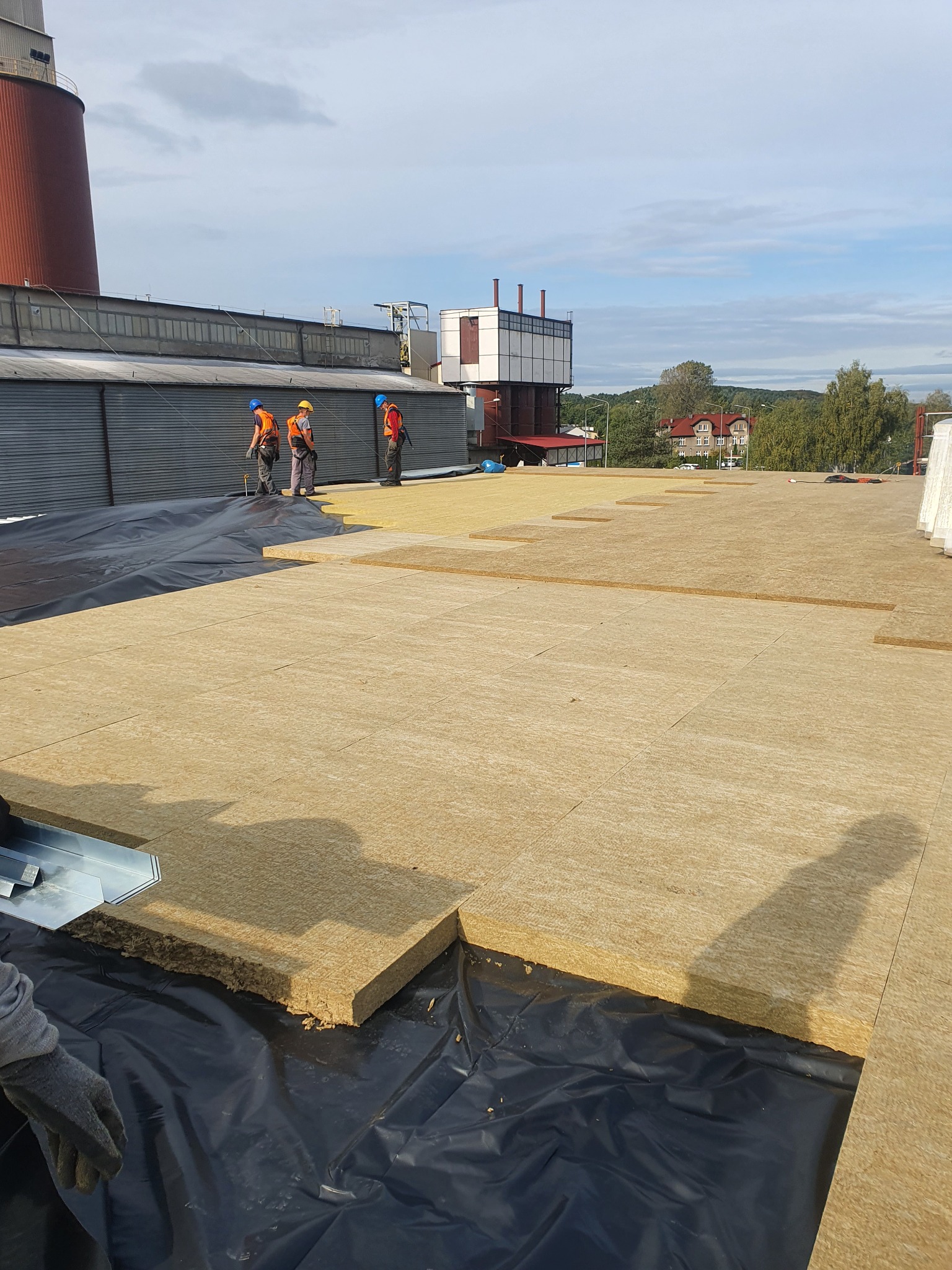 Izolacja termiczna układana na czarnej membranie dachu, pracownicy w odblaskowych kamizelkach w tle, widok na komin fabryczny i zabudowania w oddali.