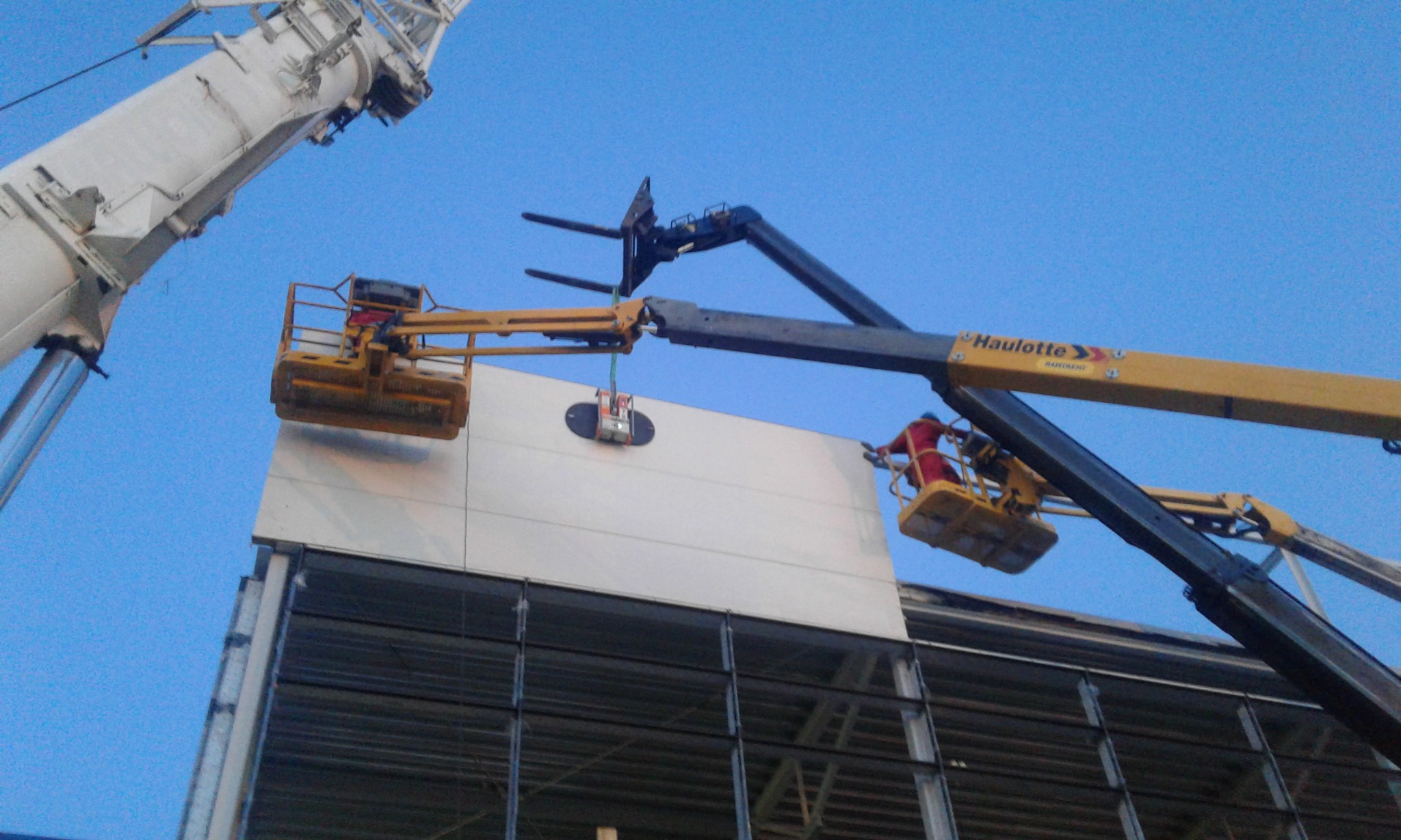 Montaż paneli elewacyjnych na wysokości z użyciem podnośników i przyssawki próżniowej, widoczny żuraw i stalowa konstrukcja budynku w tle.