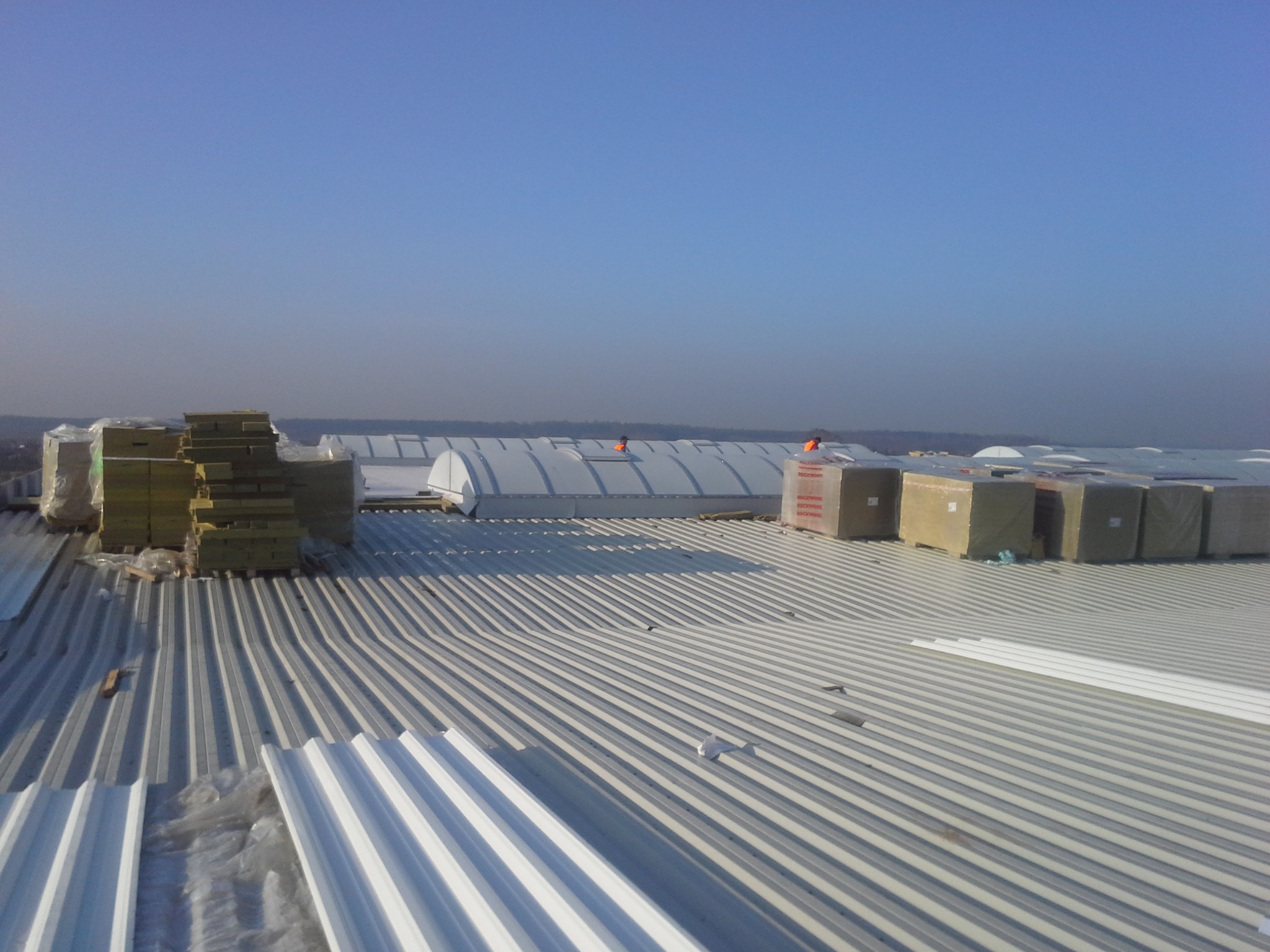 Montaż blachy trapezowej na dachu hali przemysłowej, widoczne paczki izolacji Rockwool i pracownicy w pomarańczowych kamizelkach