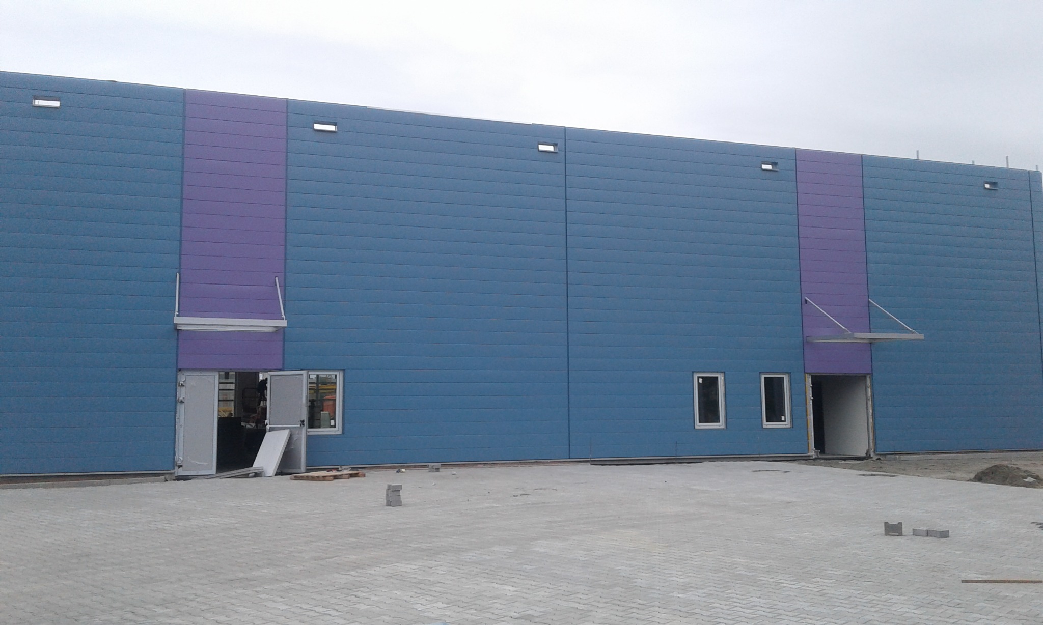 Niebiesko-fioletowa hala magazynowa z otwartymi drzwiami i oknami, widok z zewnątrz na szarej kostce brukowej.
