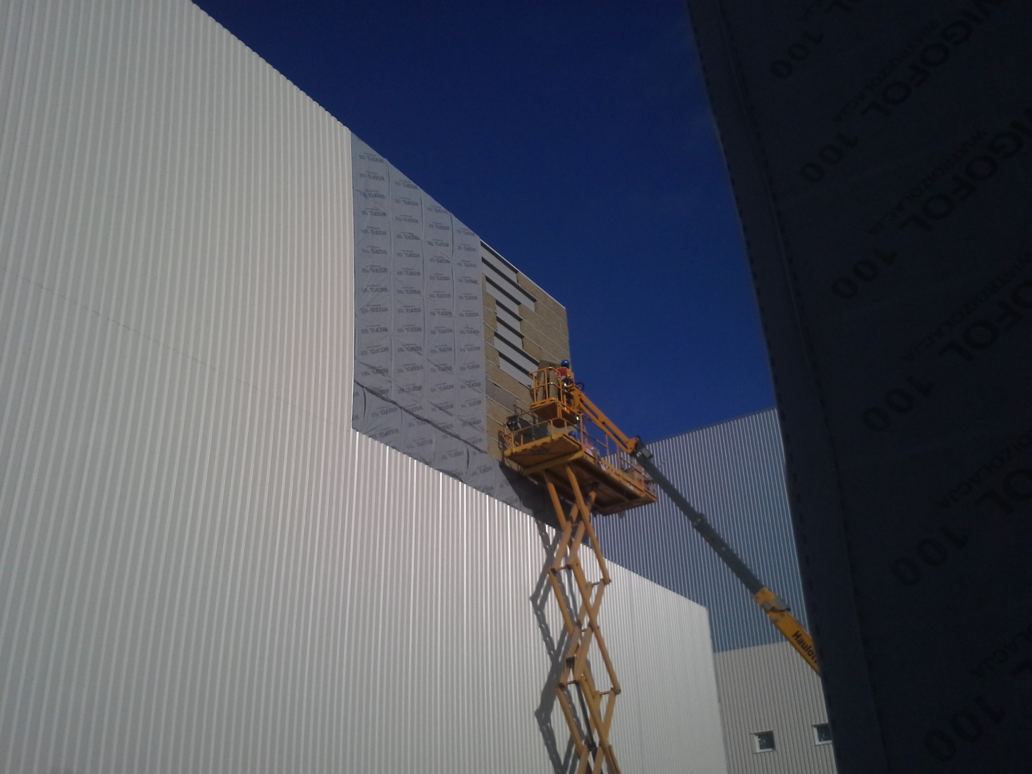 Montaż izolacji na elewacji hali przemysłowej z użyciem podnośnika koszowego, widoczne fragmenty materiału izolacyjnego z nadrukiem 'VIGOFOL 100'.