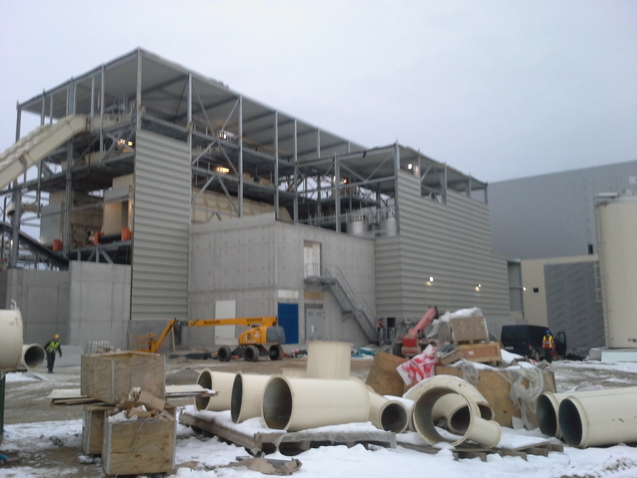 Budowa hali przemysłowej w trakcie montażu dachu, widoczne elementy konstrukcyjne, dźwigi i rury wentylacyjne na śniegu.