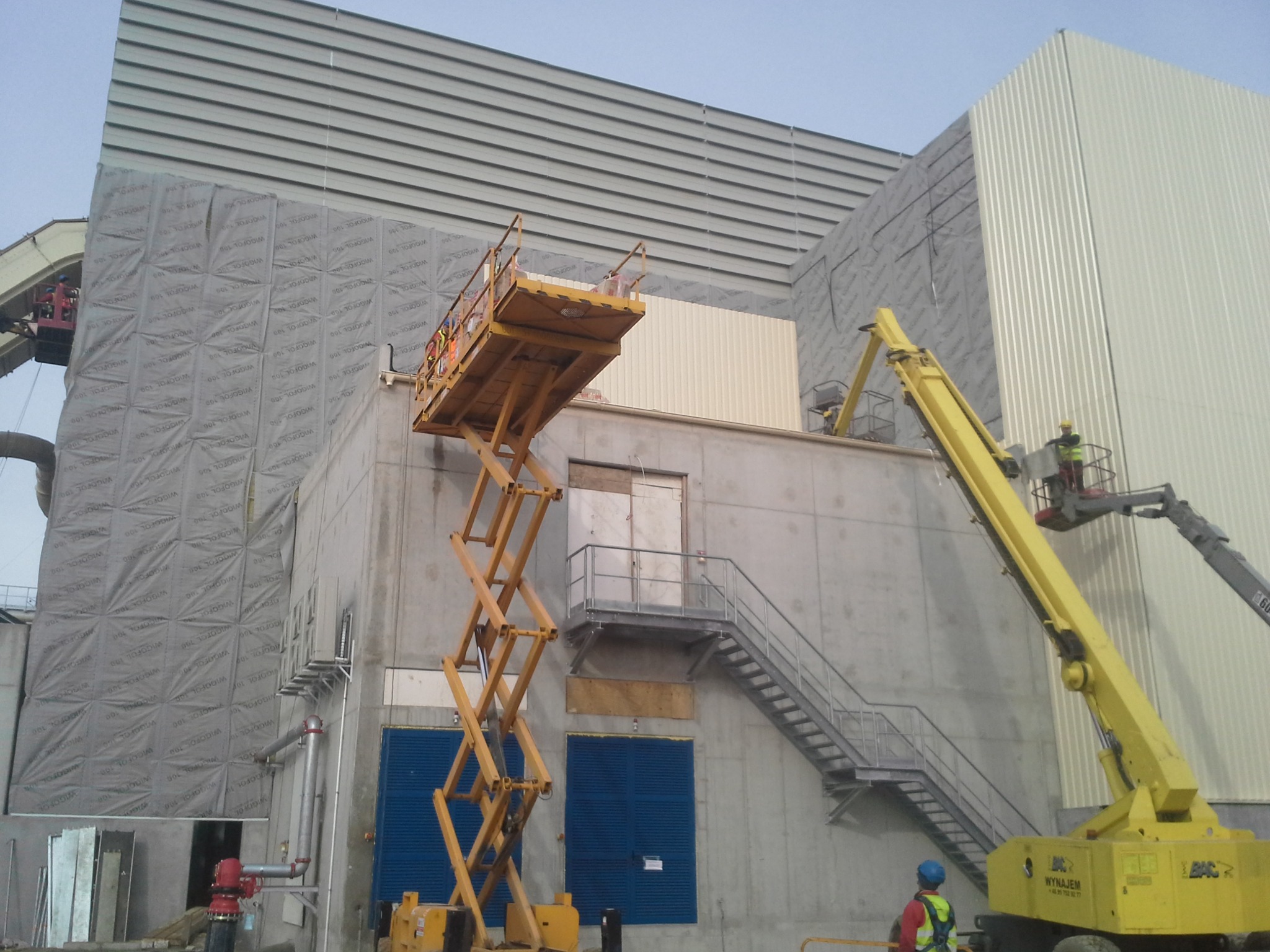 Montaż izolacji na dużej hali przemysłowej, widoczne podnośniki koszowe i pracownicy w kaskach, ściany pokryte szarą folią izolacyjną z nadrukiem Wigofol 100, w tle blacha trapezowa.