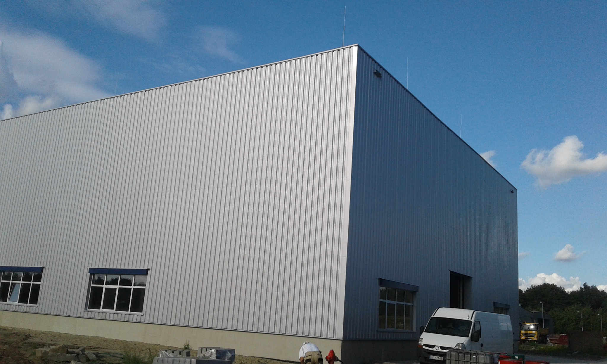Duży, srebrny budynek przemysłowy z blachy trapezowej, z widocznymi oknami, zaparkowanym białym busem i pracującymi osobami na zewnątrz, pod błękitnym, lekko zachmurzonym niebem.