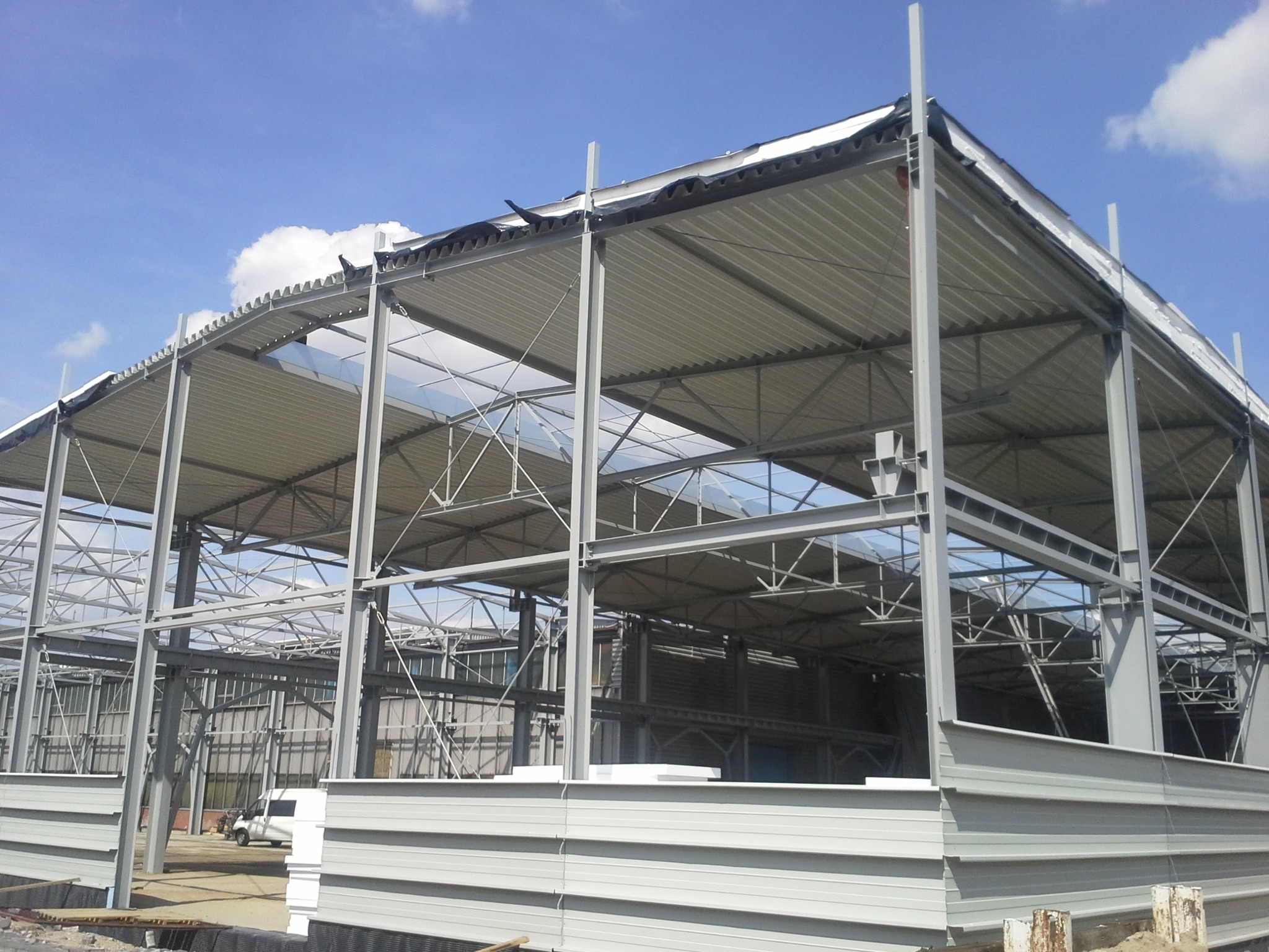 Konstrukcja stalowa hali z częściowo zamontowanym dachem z blachy trapezowej, widoczne elementy mocujące i membrany dachowe, na tle błękitnego nieba z chmurami.
