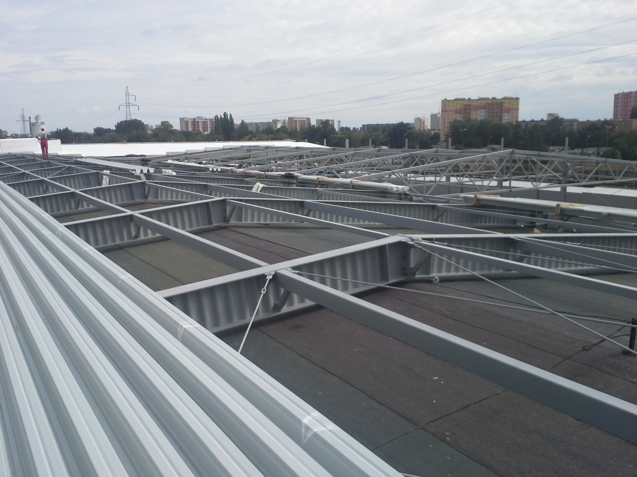 Konstrukcja stalowa dachu płaskiego z widocznym pracownikiem w oddali, na tle zabudowy miejskiej i pochmurnego nieba.