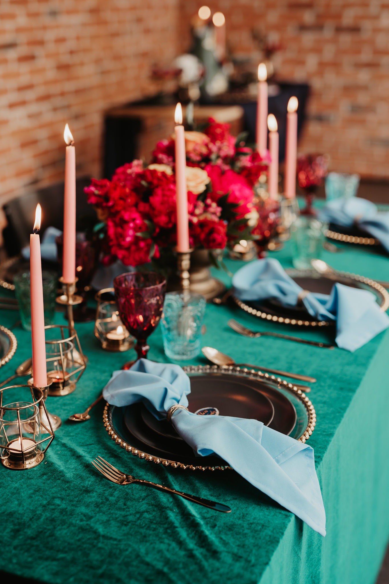 Stylowa aranżacja stołu z turkusowym obrusem, złotą zastawą, pudrowymi świecami w geometrycznych świecznikach i bukietem czerwonych kwiatów, uzupełniona o burgundowe kieliszki.