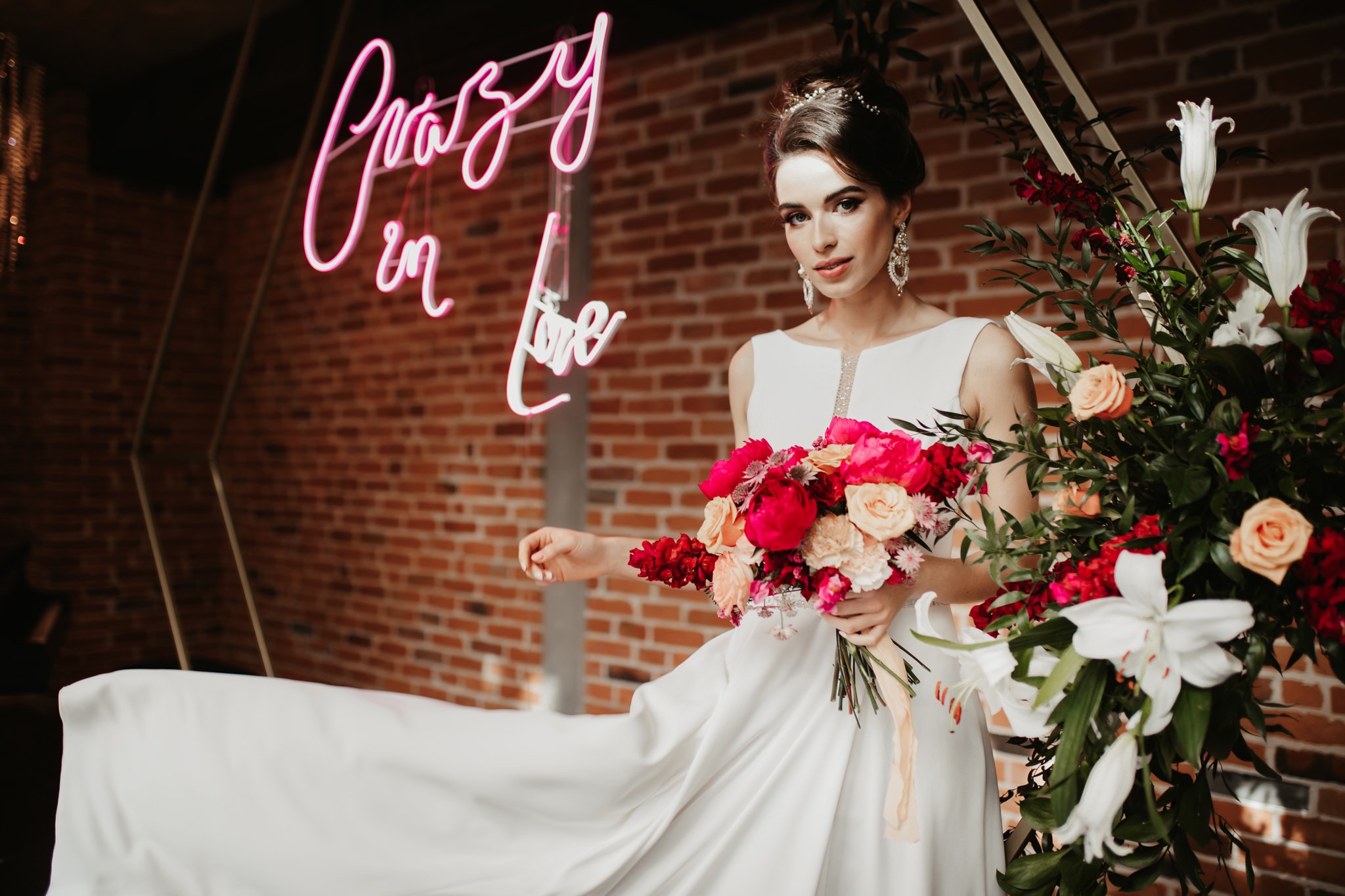 Panna młoda w białej sukni trzyma bukiet różowych i brzoskwiniowych kwiatów na tle ceglanej ściany z neonowym napisem 'Crazy in Love'.