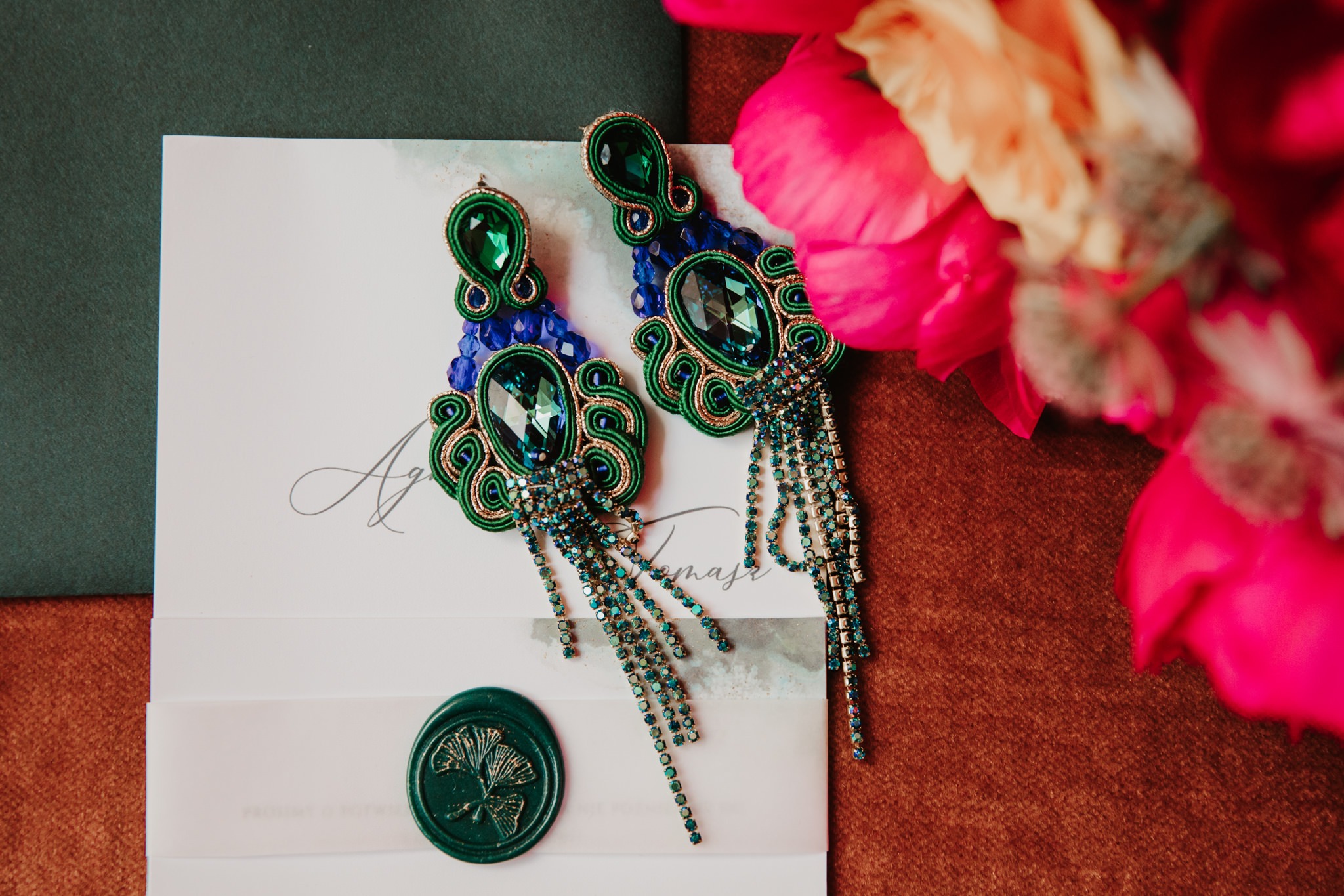 Dwa kolczyki sutasz z zielonymi i niebieskimi kryształami, leżące na zaproszeniu ślubnym z zieloną lakową pieczęcią, obok różowych kwiatów.