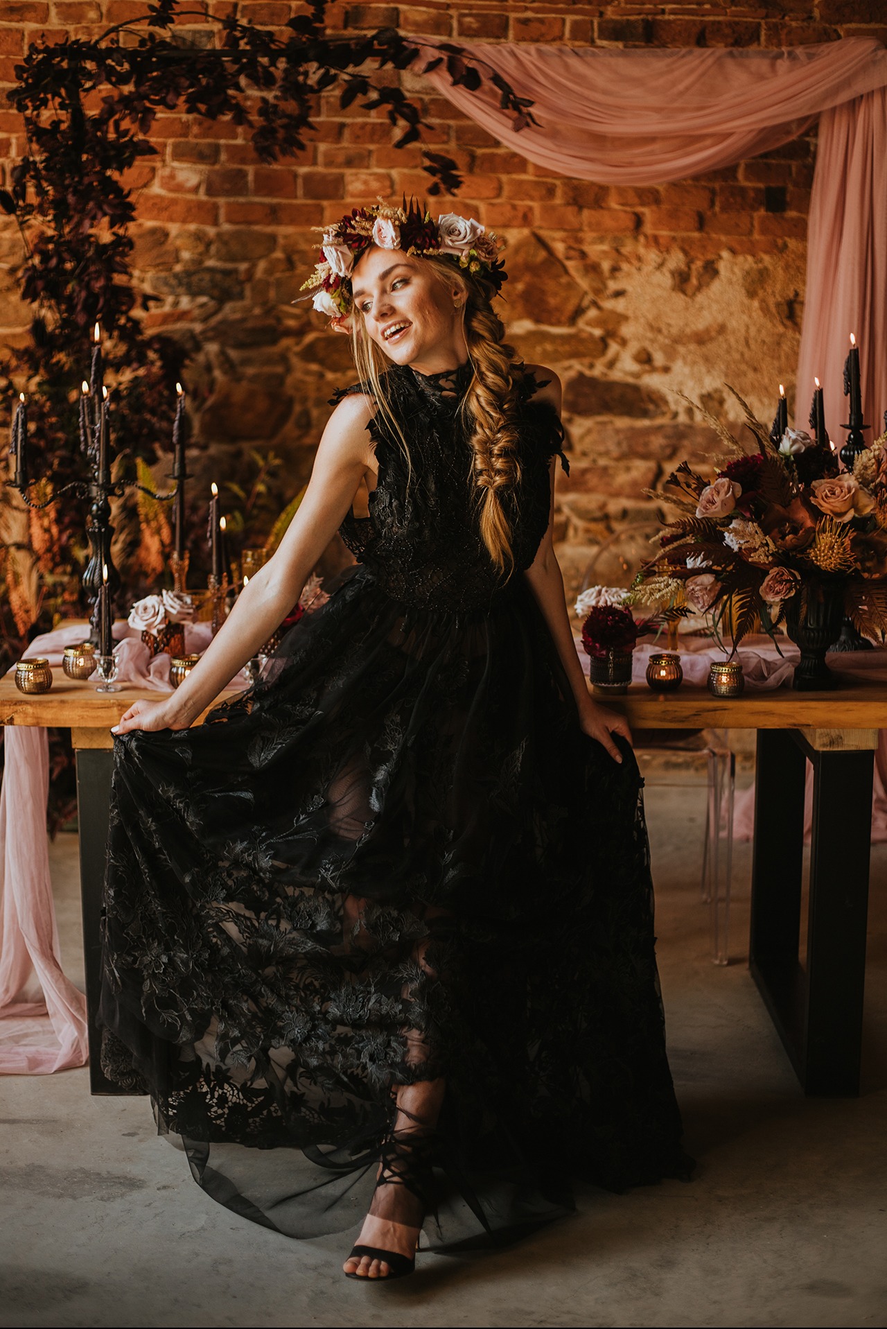 Stylizowana sesja zdjęciowa: kobieta w czarnej, koronkowej sukni i wianku z kwiatów, pozująca przy stole z dekoracjami w mrocznym zamku z ceglanym tłem.