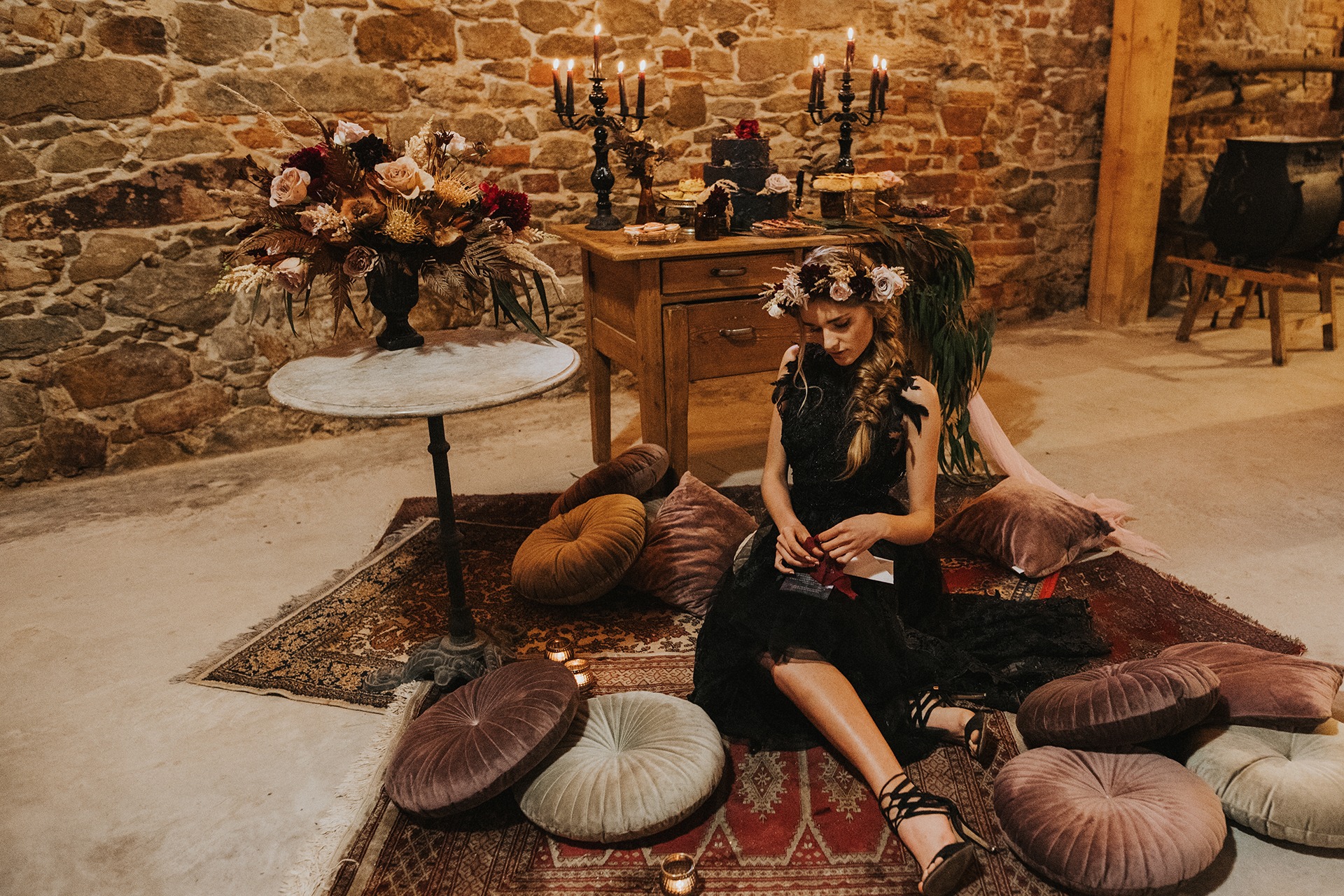 Stylizowana sesja w mrocznym zamku: modelka w czarnej sukni siedzi na dywanie otoczona poduszkami, w tle kamienna ściana, stół z dekoracjami i świecami.