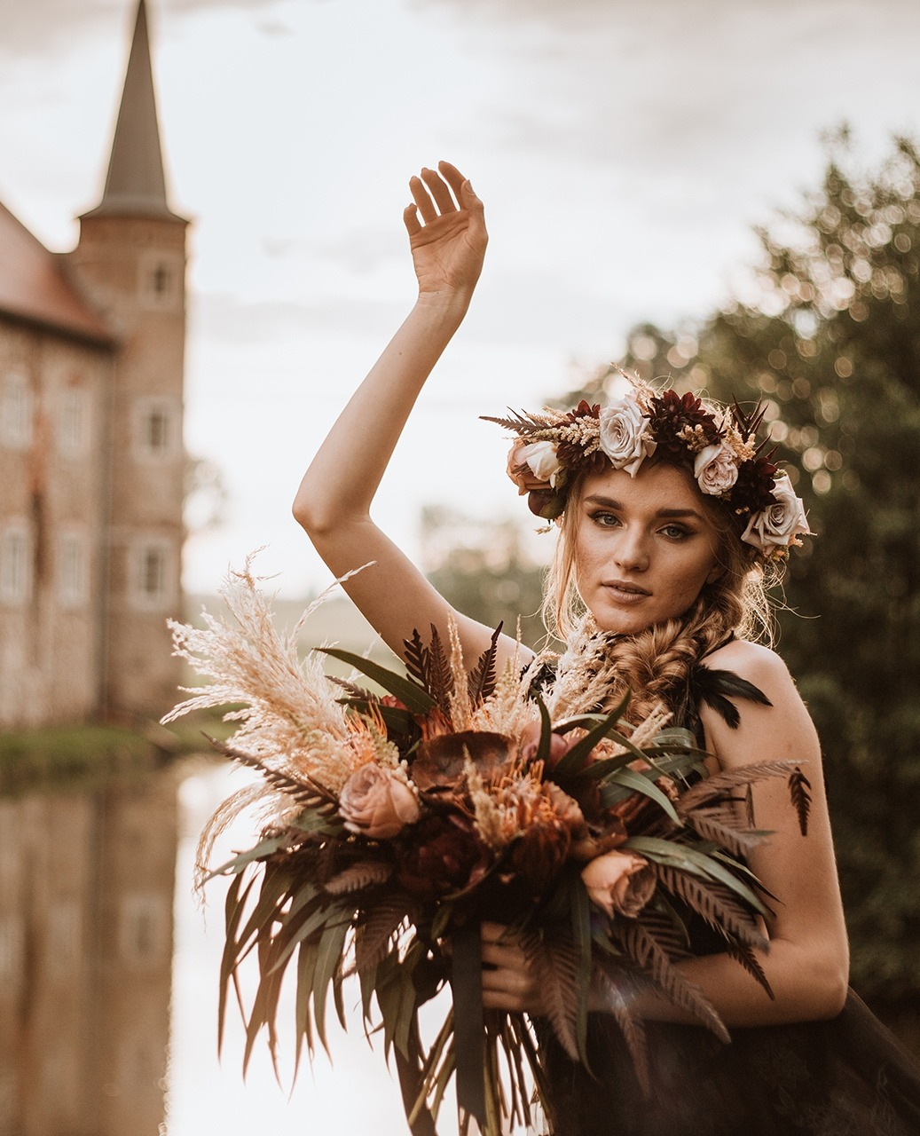 Modelka w koronie z kwiatów pozuje z bukietem w stylu rustykalnym na tle zamku i jeziora, sesja stylizowana.