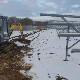 Żółta minikoparka w zimowym krajobrazie, przygotowująca teren pod konstrukcję stalową, z widocznymi śladami opon na śniegu i wykopaną ziemią.