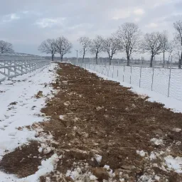 Przygotowany pas ziemi pod instalację ogrodzenia panelowego na zimowym terenie, z widocznymi elementami konstrukcji pod panele solarne w tle.