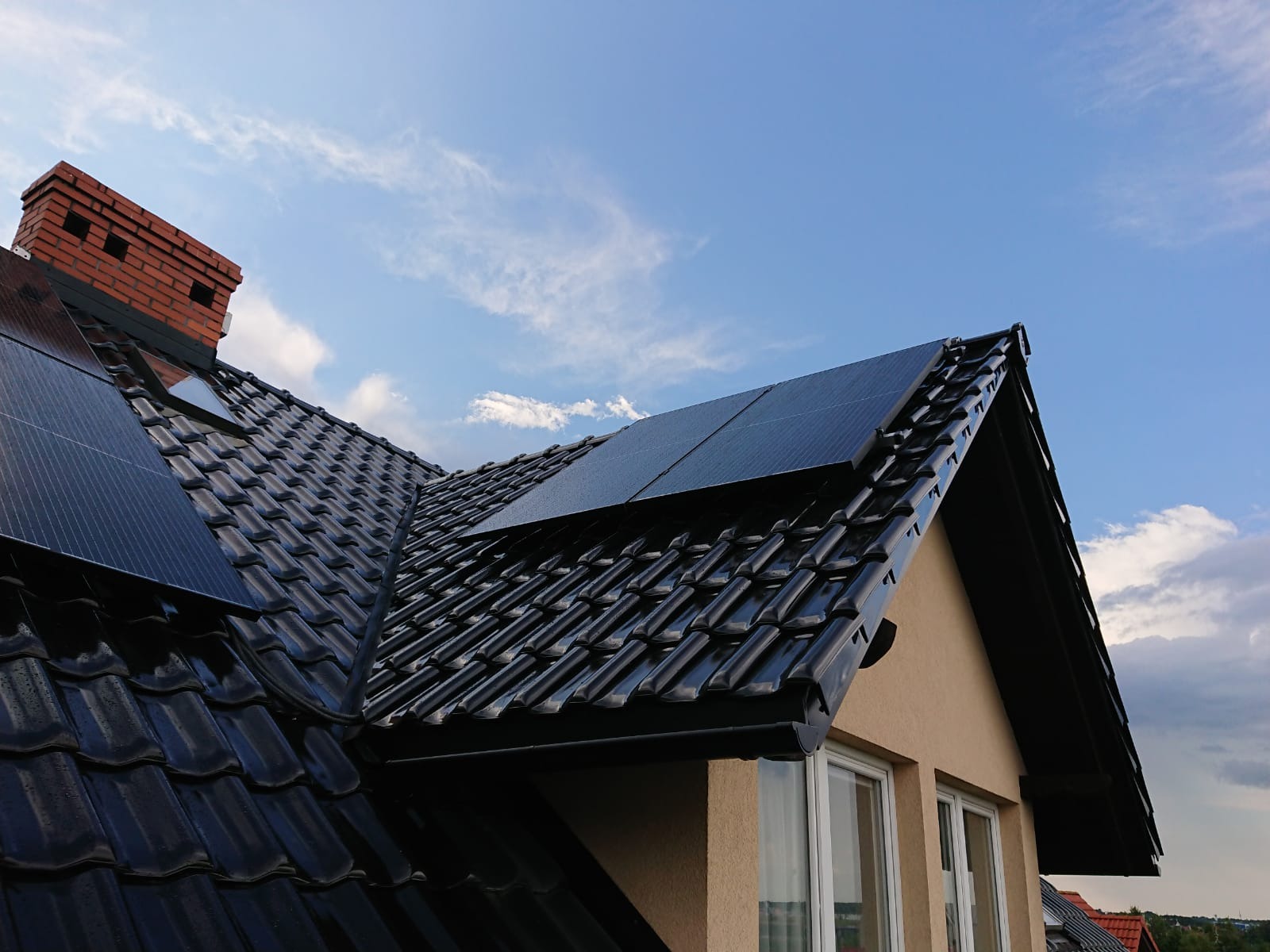 Panele słoneczne na błyszczącym, czarnym dachu z dachówki ceramicznej w Gdańsku, widok z dołu na tle błękitnego nieba z chmurami.