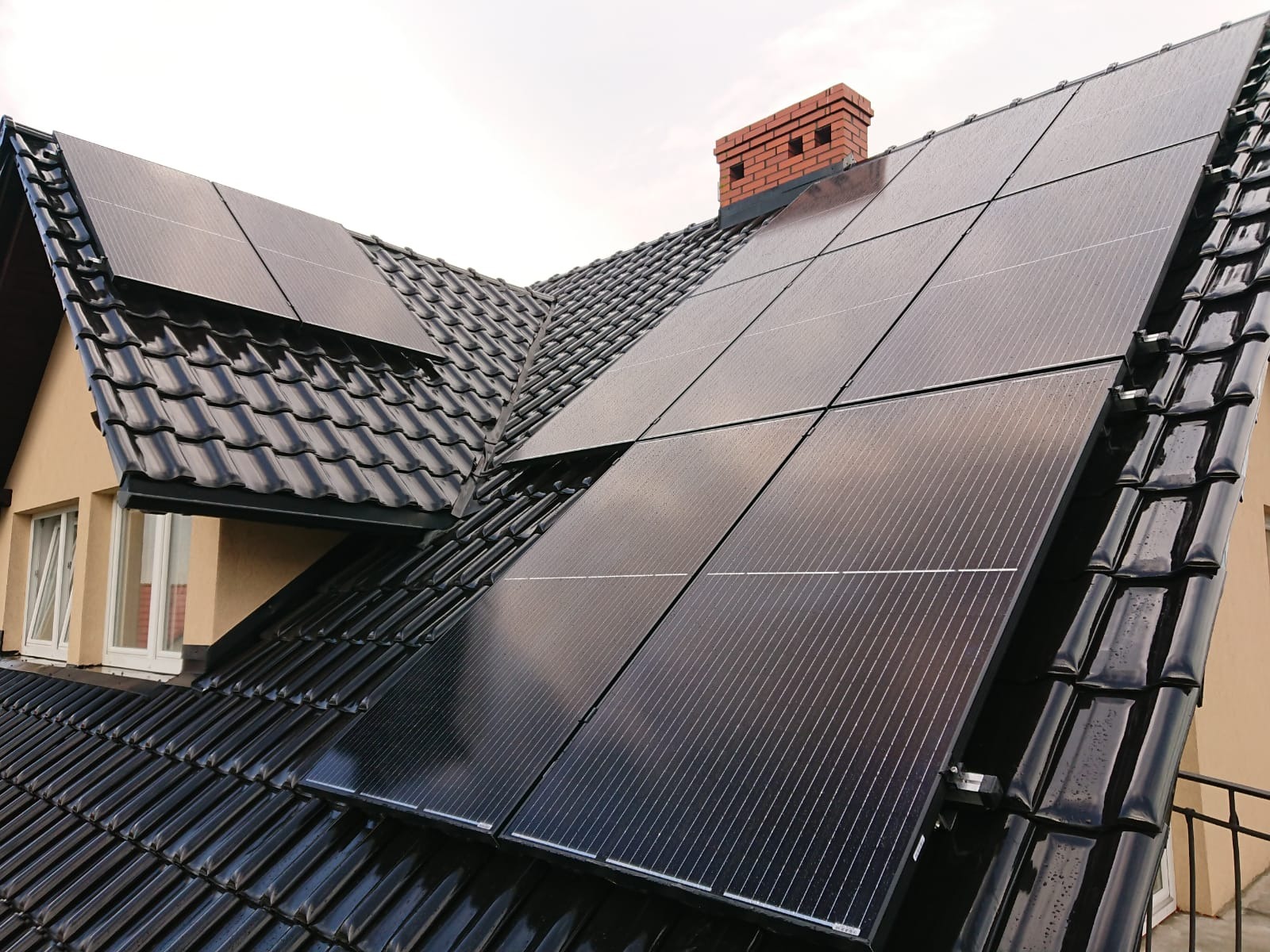 Czarne panele słoneczne zamontowane na błyszczącym, czarnym dachu z dachówki ceramicznej, widok na komin z cegły w tle.