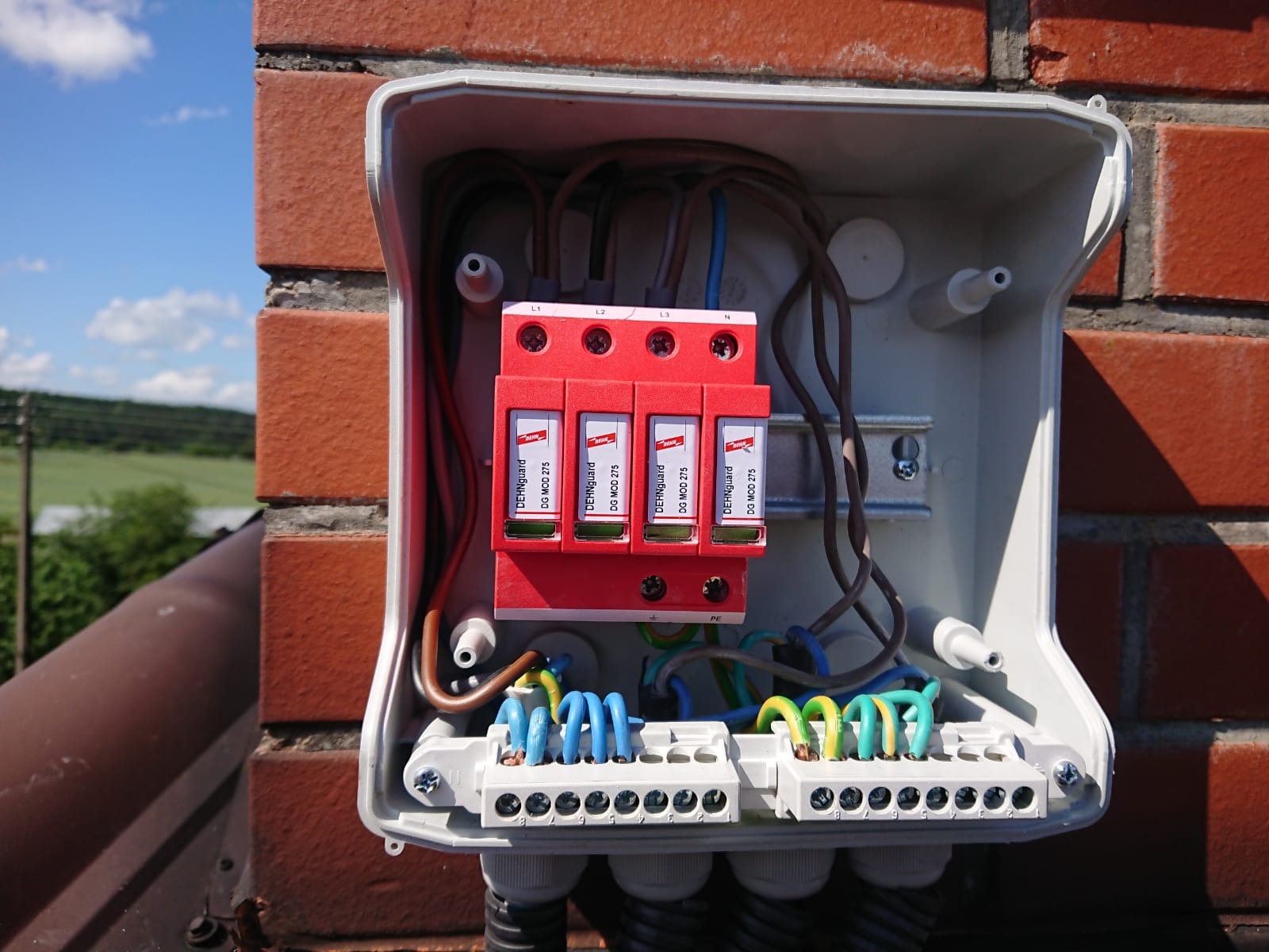 Otwarta skrzynka z zabezpieczeniem przeciwprzepięciowym DehnGuard DG MOD 275, zamontowana na ceglanym kominie z widocznymi przewodami w kolorach brązowym, niebieskim i zielono-żółtym.
