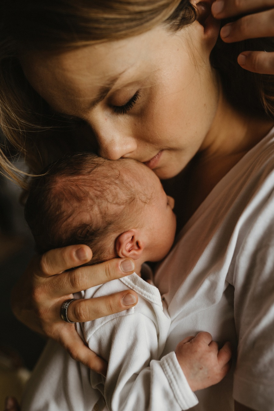 Delikatne ujęcie matki całującej w głowę noworodka, z bliska widoczna faktura włosów dziecka i pierścionek na palcu matki.
