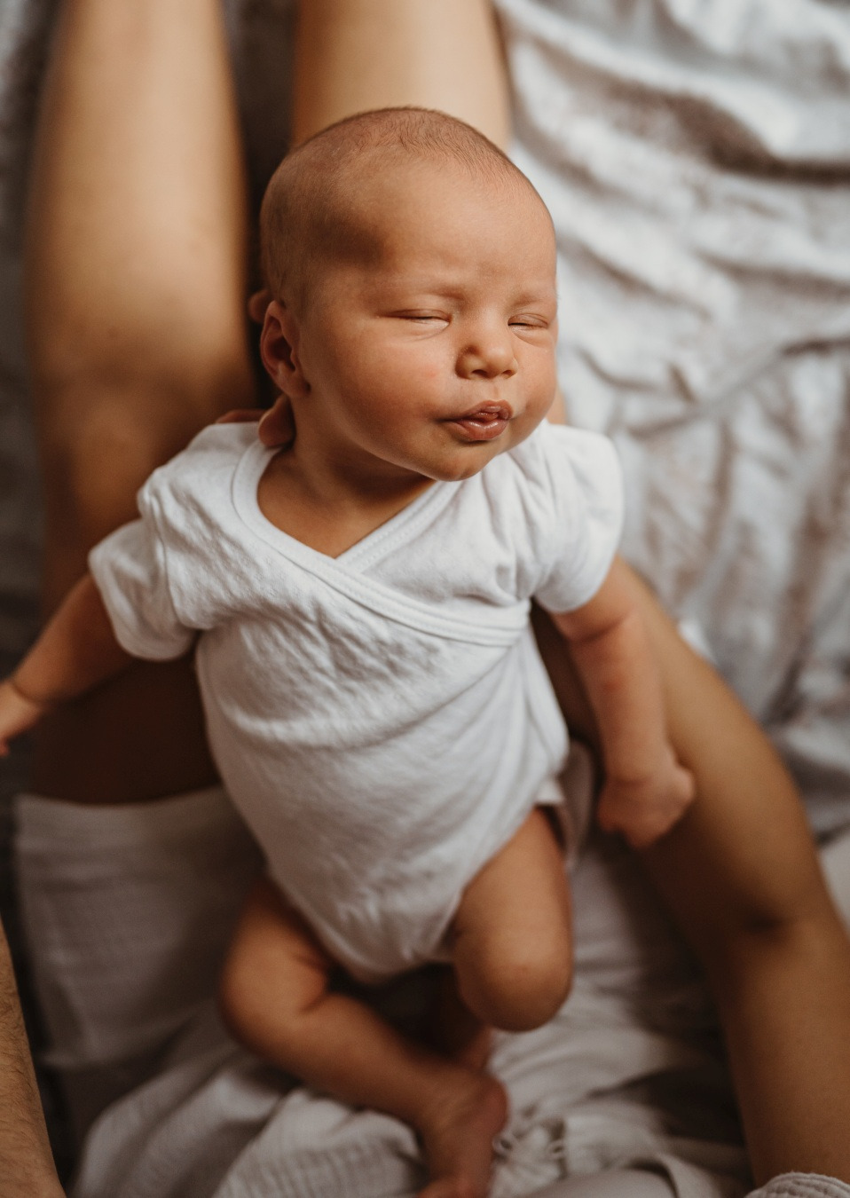 Śpiący noworodek w białym body, trzymany na kolanach osoby dorosłej, delikatne światło, neutralne tło.