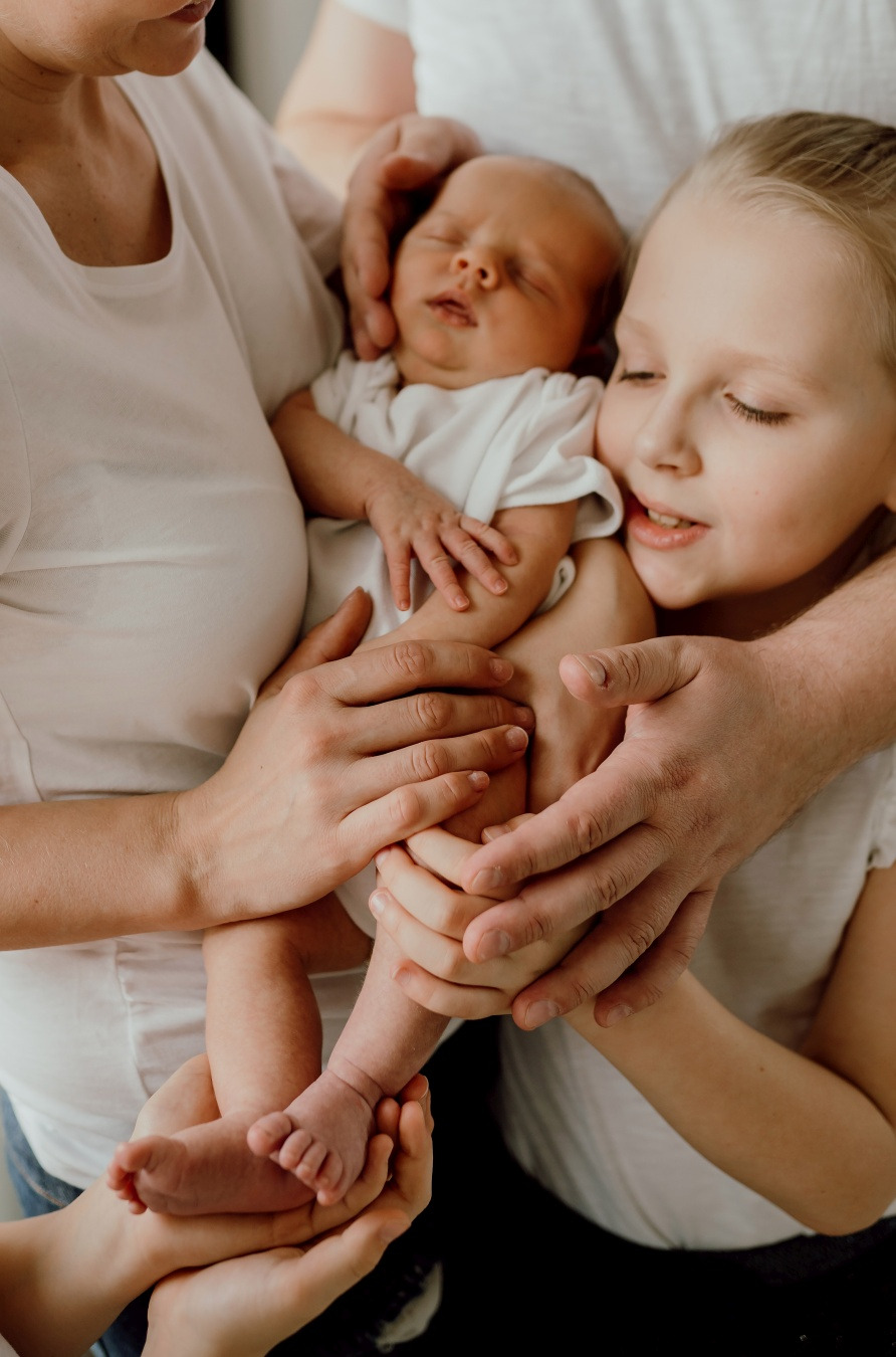 Rodzinne ujęcie: śpiący noworodek w ramionach rodziców i starszej siostry, wszyscy ubrani w białe koszulki, naturalne światło.
