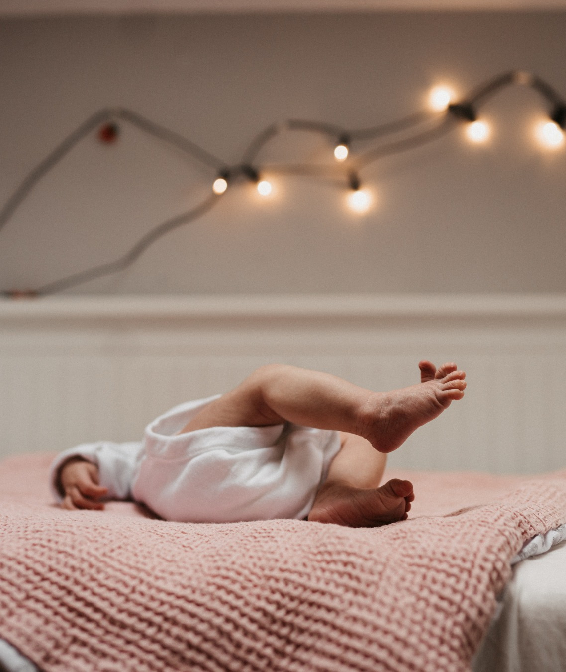 Nóżki noworodka w białym ubranku leżą na różowym kocu w pokoju z lampkami w tle.