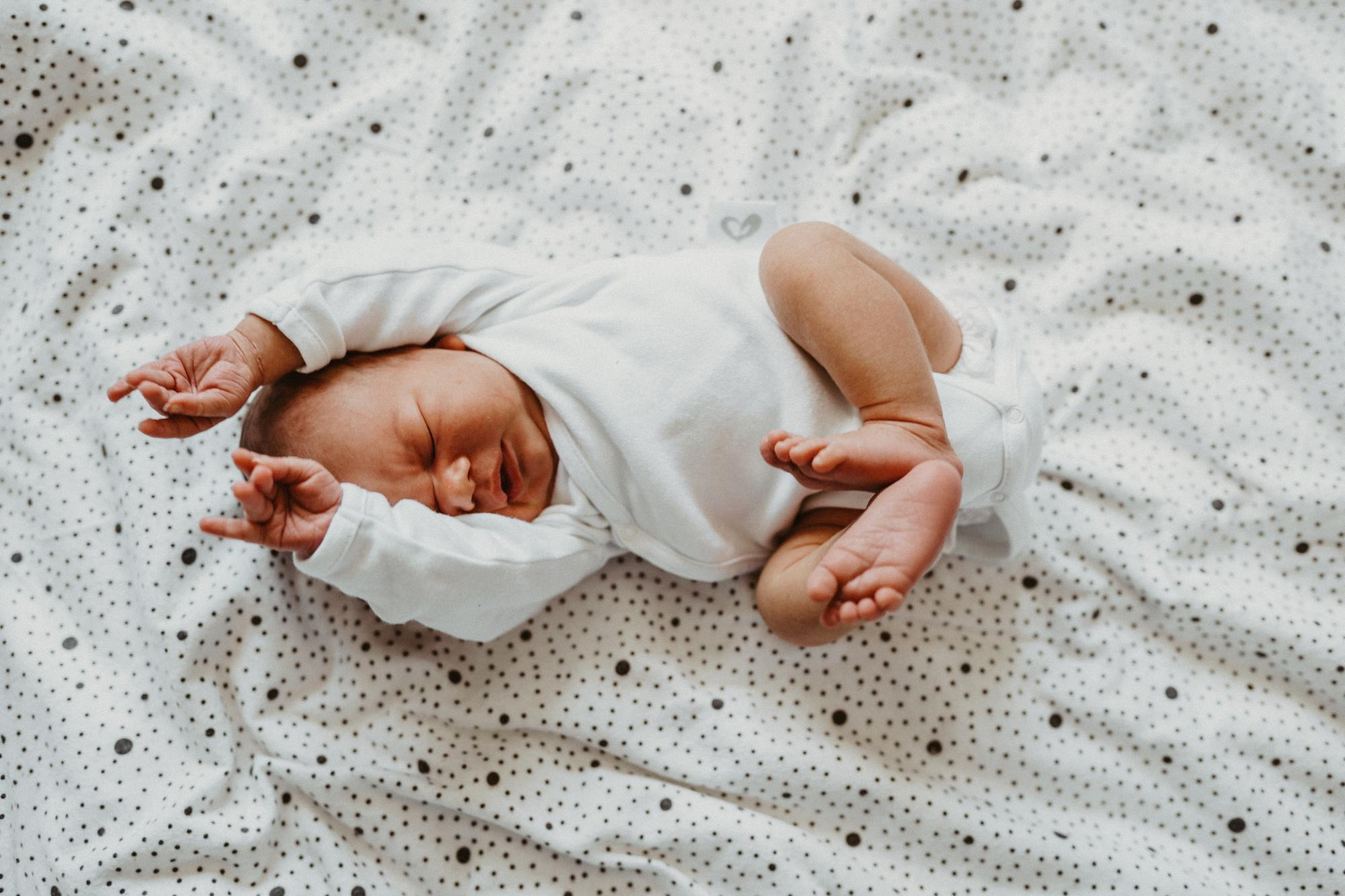 Śpiący noworodek w białym body leży na białym kocyku w czarne kropki, z rączkami uniesionymi do góry, widoczne małe stópki.