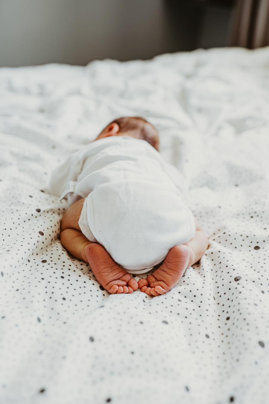 Noworodek leży na brzuszku na łóżku w białym body, widoczne małe stópki i plecki.