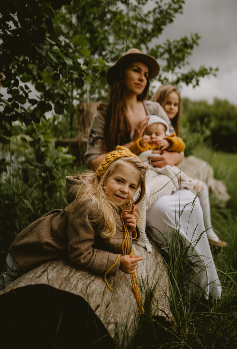 Matka z trzema córkami siedząca na powalonym drzewie w plenerze, najmłodsza córka w żółtym swetrze na kolanach matki, najstarsza córka w brązowej kurtce leży na drzewie i patrzy w obiektyw, a w tle...