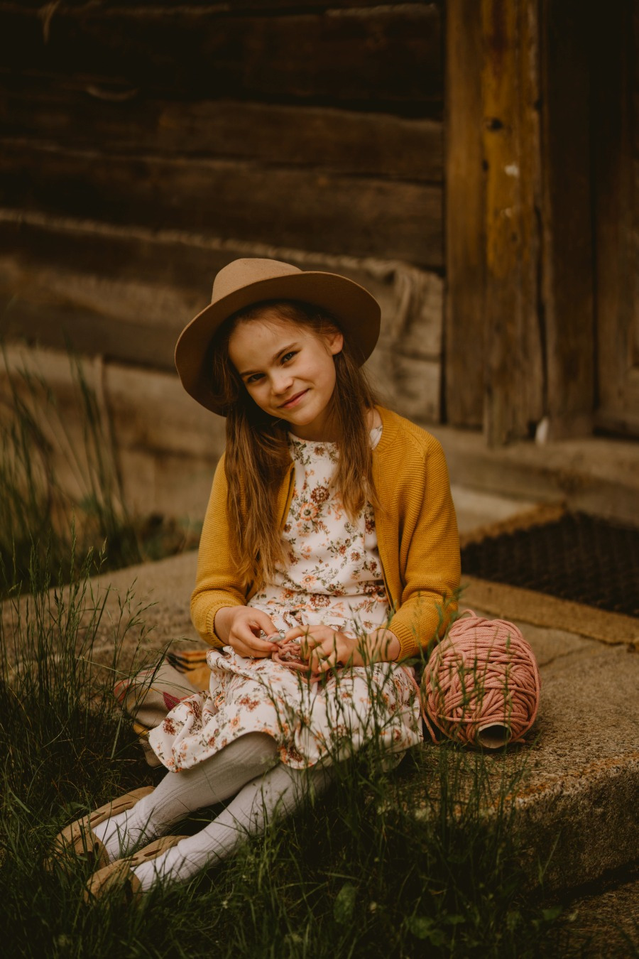 Dziewczynka w kapeluszu i kwiecistej sukience siedzi na schodach drewnianego domu, bawiąc się różowym sznurkiem do makramy.