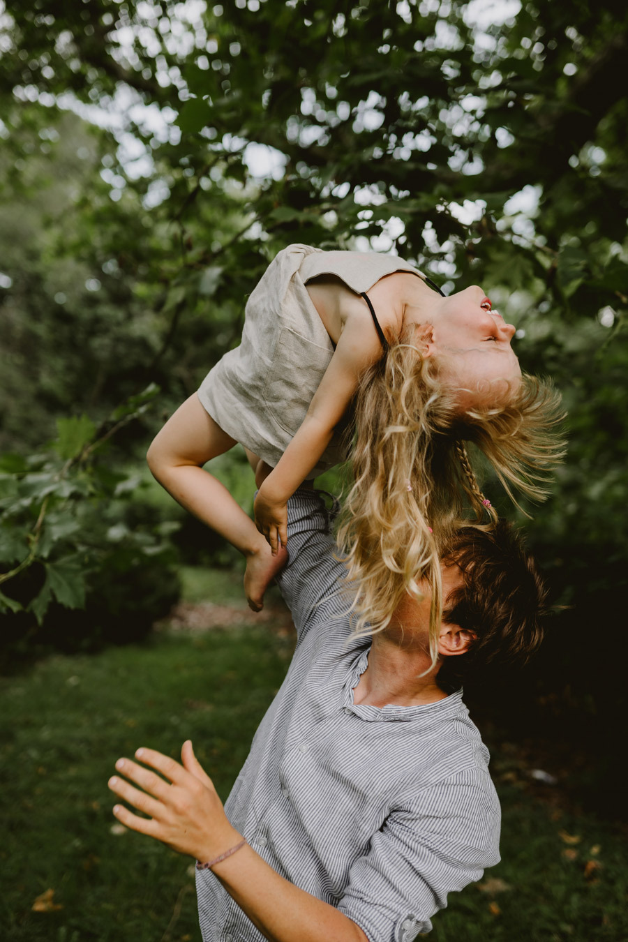 Ojciec trzymający córkę do góry nogami, śmiejąca się dziewczynka w lnianej sukience, plener w zieleni.