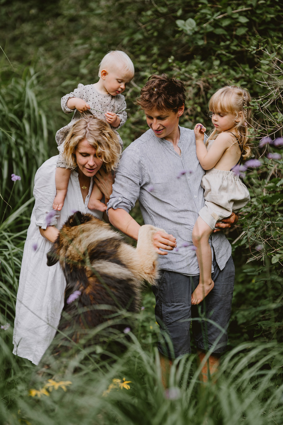 Rodzina z dwójką małych dzieci i dużym psem w plenerze, matka trzyma na ramionach niemowlę, ojciec trzyma na rękach starszą dziewczynkę, w tle zielona roślinność.