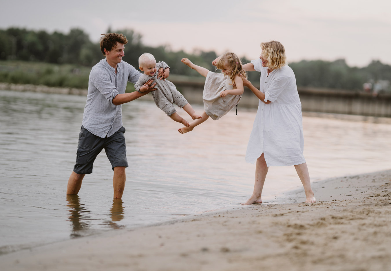 Rodzice podrzucają w górę dwójkę uśmiechniętych dzieci na piaszczystej plaży nad brzegiem rzeki; tata stoi w wodzie, mama na piasku.