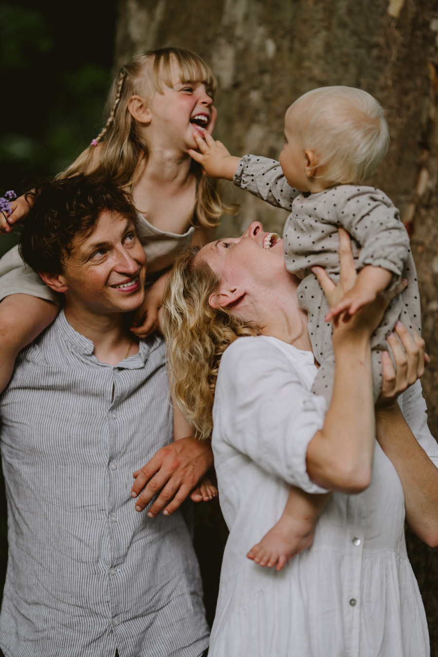 Radosna sesja rodzinna w plenerze: tata z córką na barana, mama trzymająca niemowlę, wszyscy śmieją się na tle drzewa.