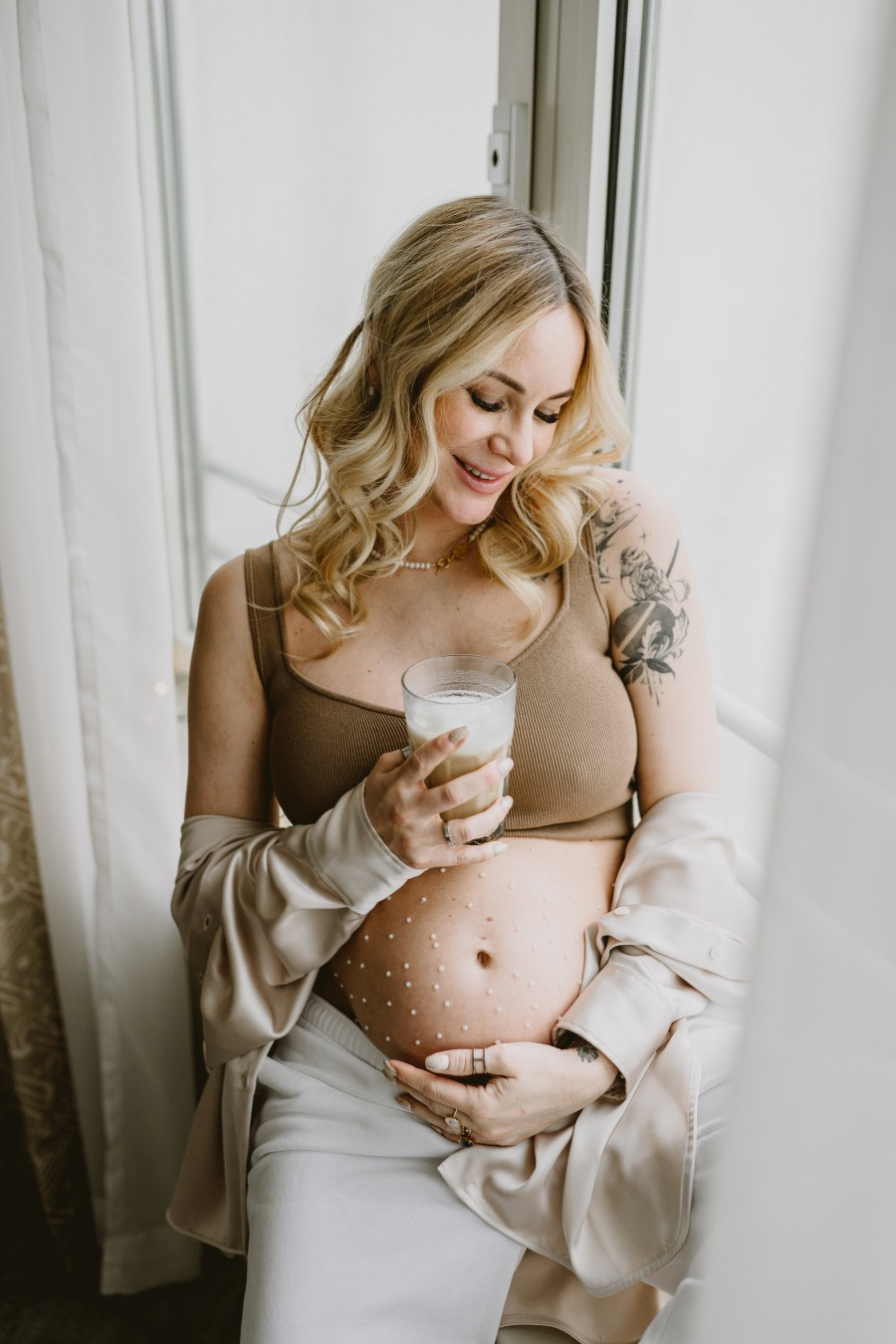 Kobieta w ciąży, z blond włosami i tatuażem na ramieniu, trzyma szklankę z napojem, na brzuchu naklejone białe kropki, siedzi w jasnym pomieszczeniu przy oknie.