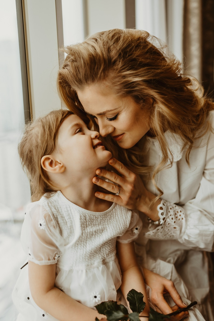 Matka z córką w białych sukienkach siedzą blisko okna, dotykając się nosami w czułym ujęciu.