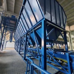 Długa linia niebieskich metalowych zbiorników na konstrukcji wsporczej w hali przemysłowej z betonowym sufitem i podłogą z kostki brukowej.