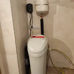 Biały zmiękczacz wody z czarnym sterownikiem i zbiornikiem wyrównawczym zamontowany na ścianie wyłożonej beżowymi płytkami.